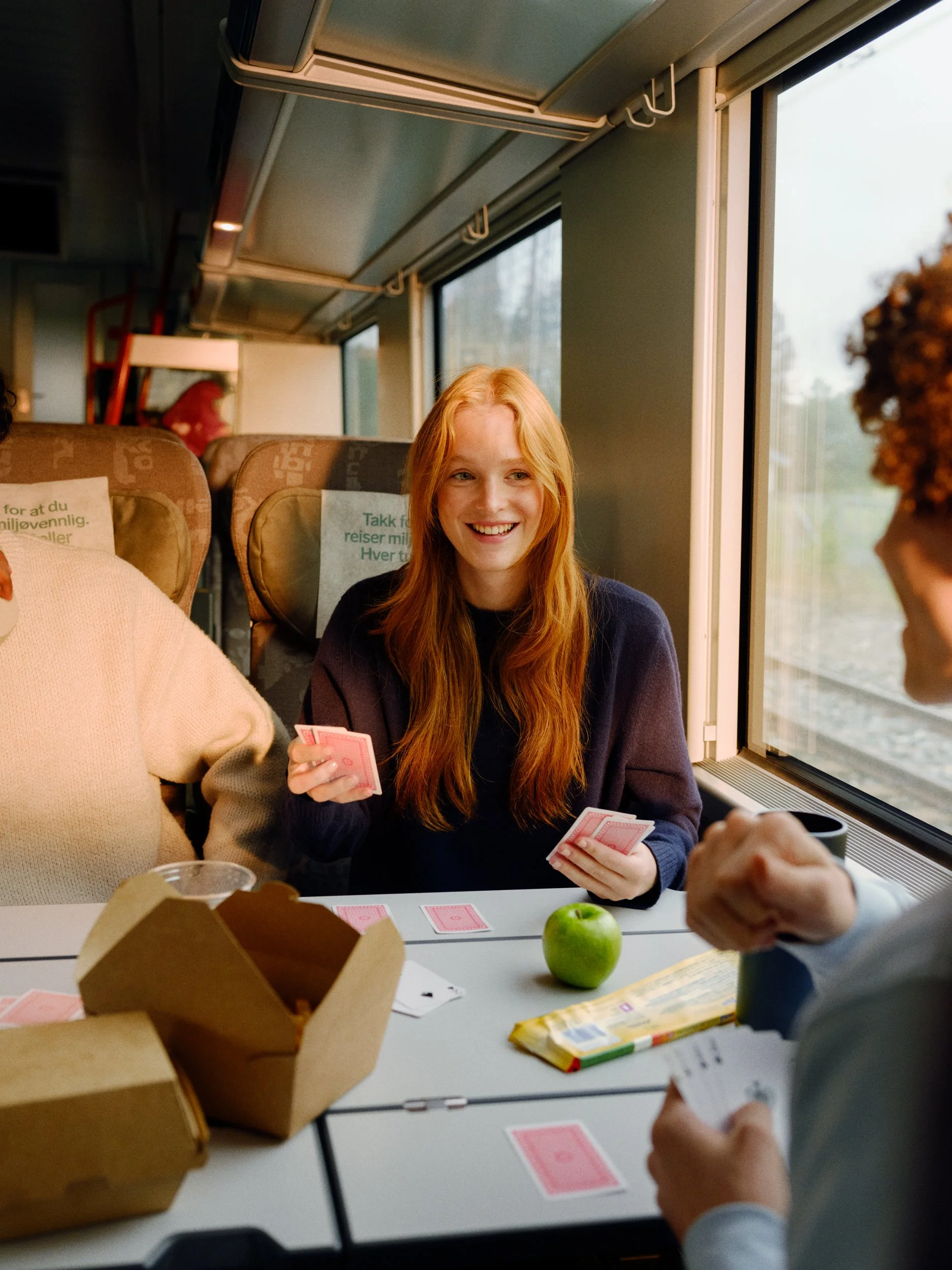 A group of young people playing cards while sitting at a table on a train. There is a green apple and some snacks on the table, and the passengers are smiling and enjoying their time.