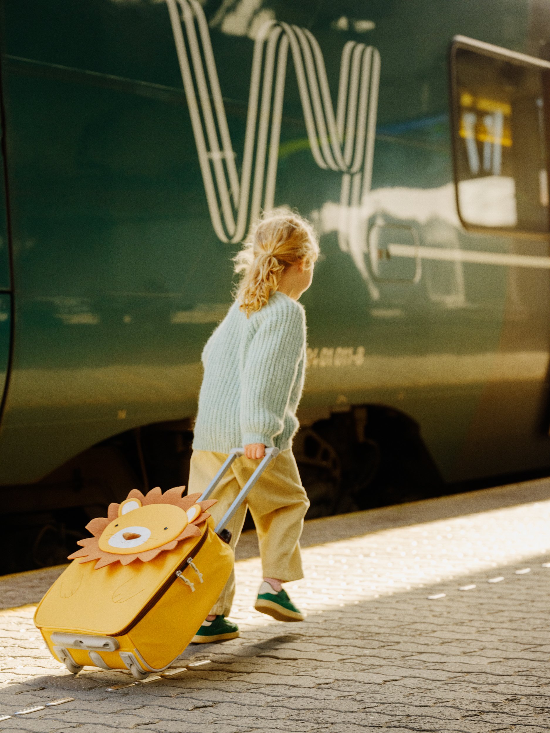 A young girl with curly blonde hair walking with a yellow suitcase that has a lion face on the front, next to a green train.