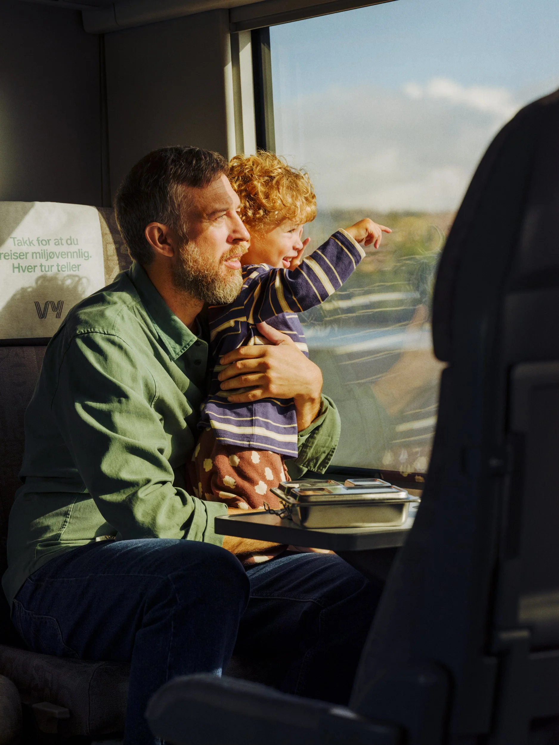 A man and a young girl sitting together on a bus, with the girl pointing out the window and the man smiling. The girl has curly red hair and is wearing a striped shirt and patterned pants. The man has a beard and is wearing a green shirt. There is a 
