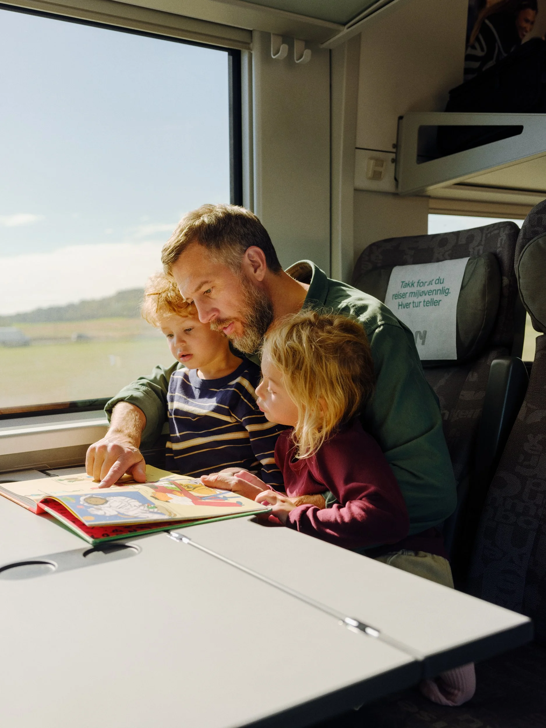 A man with two children reading a book together on a train, with a window showing a rural landscape outside.