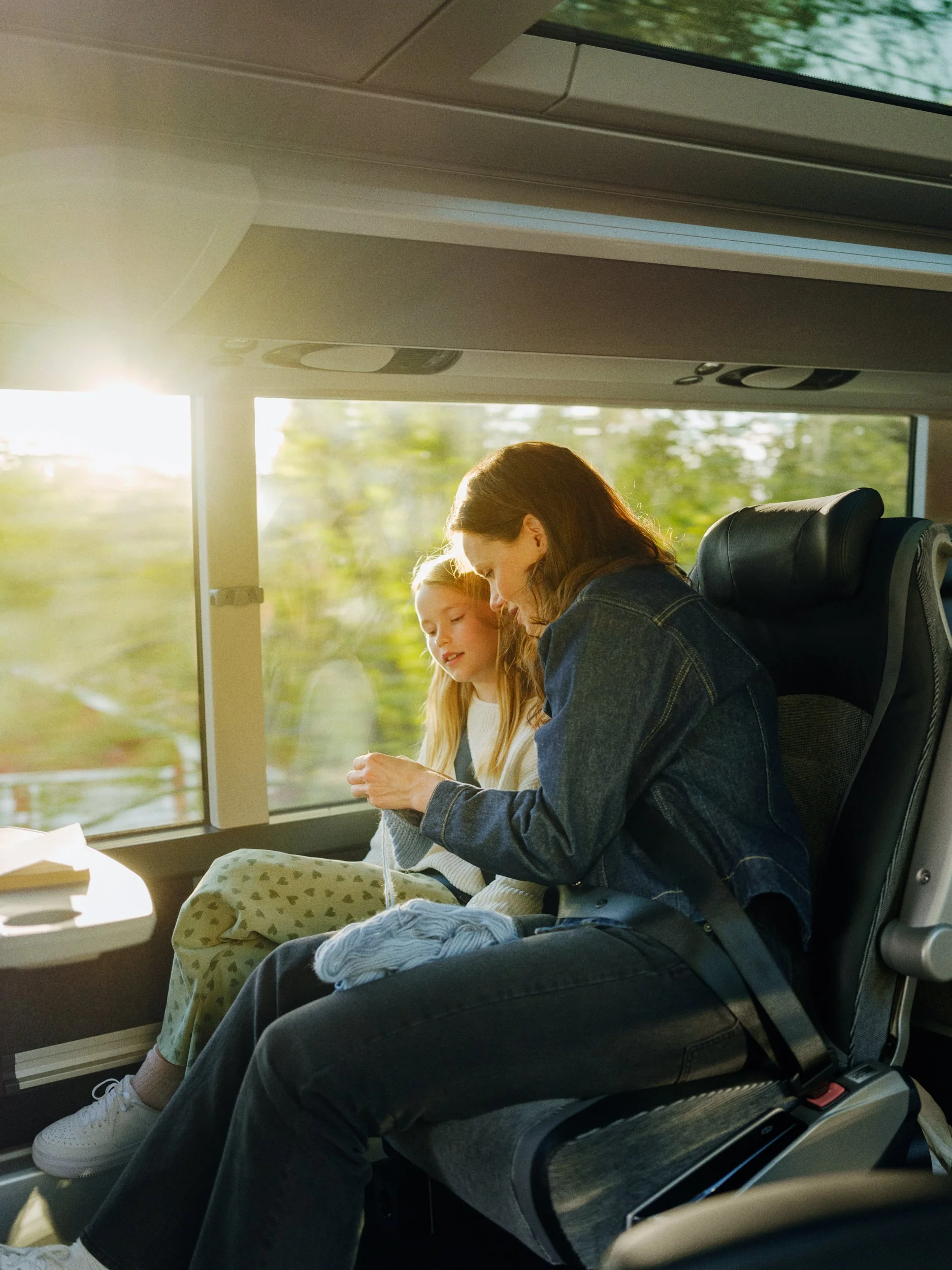 A woman and girl sitting on a bus, knitting and chatting while enjoying sunlight through the window.