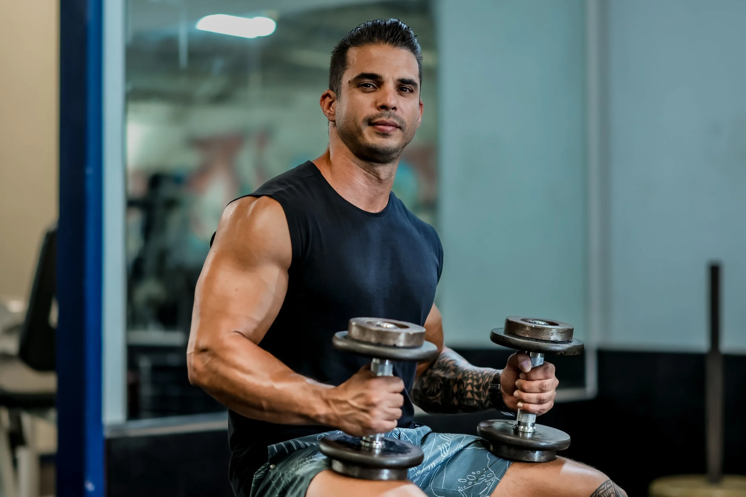 A picture of Daniel wearing a black sleeveless shirt, working out in a gym, lifting dumbbells.