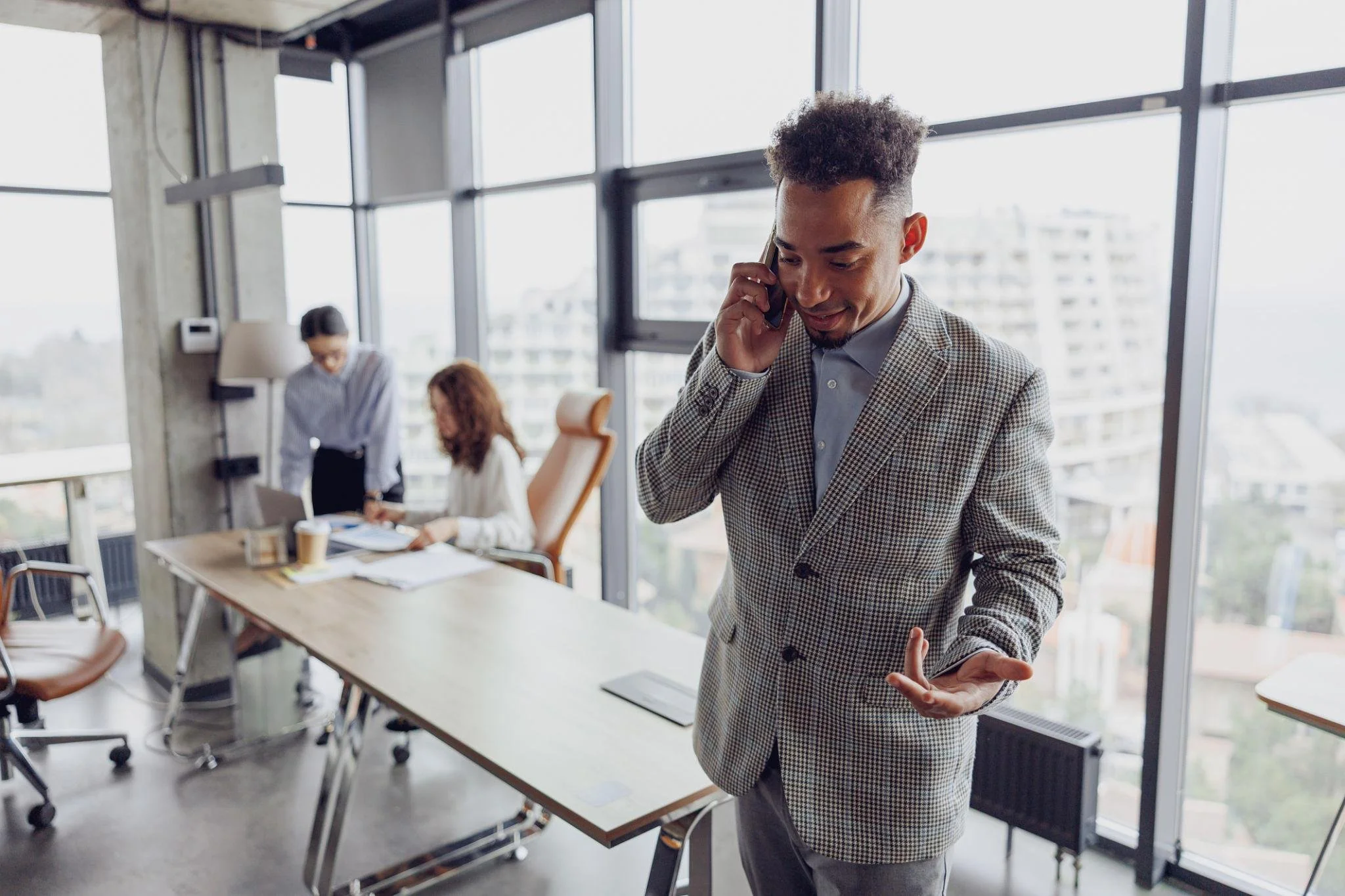 A man in a checked blazer talking on a mobile phone in a modern office meeting room with large windows, a long table, and people working at the table in the background.