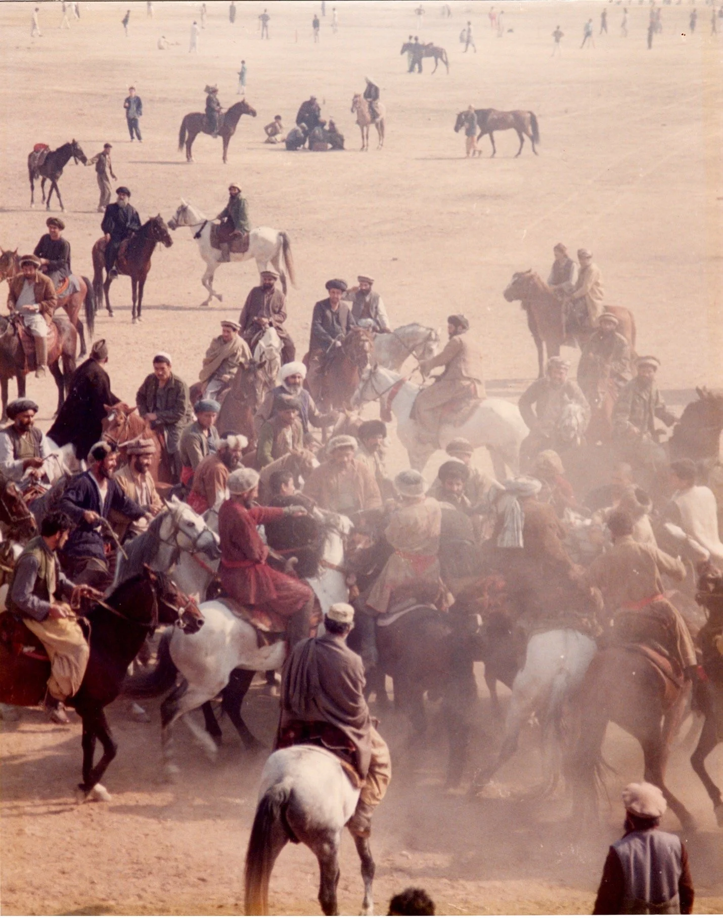 A large gathering of people and horses in a desert or dusty area, with some people riding horses and others standing or sitting nearby.
