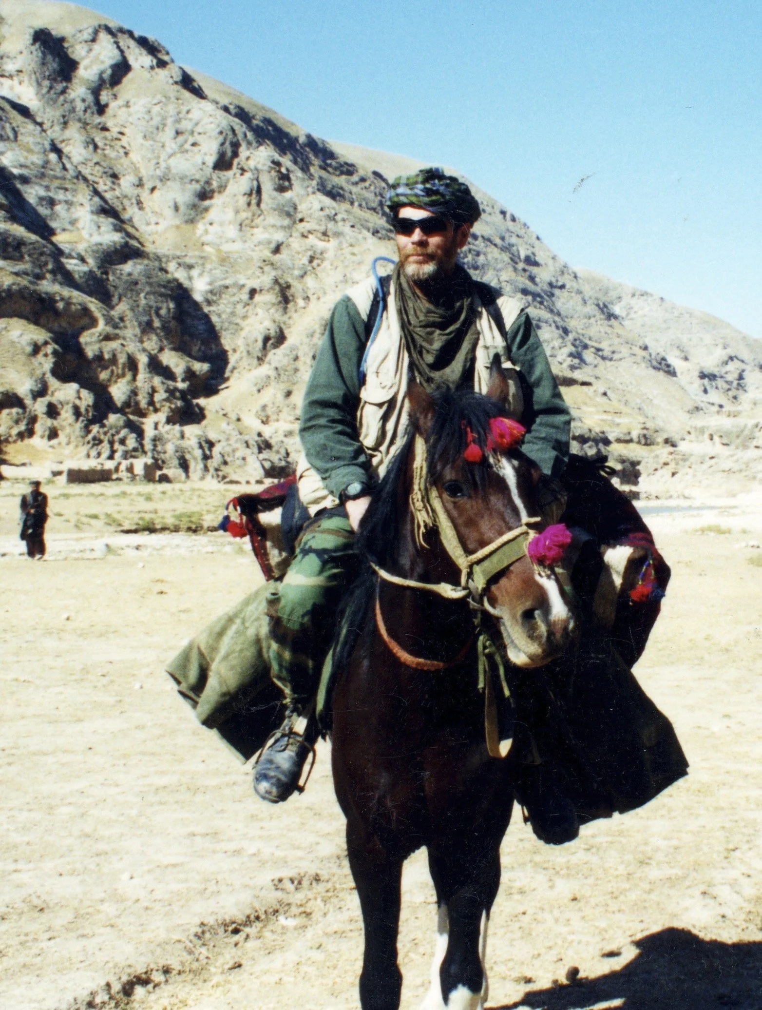 A man riding a horse in a desert landscape with large rocky mountains in the background. He is wearing sunglasses, a green jacket, and a beige vest, and has a scarf around his neck.