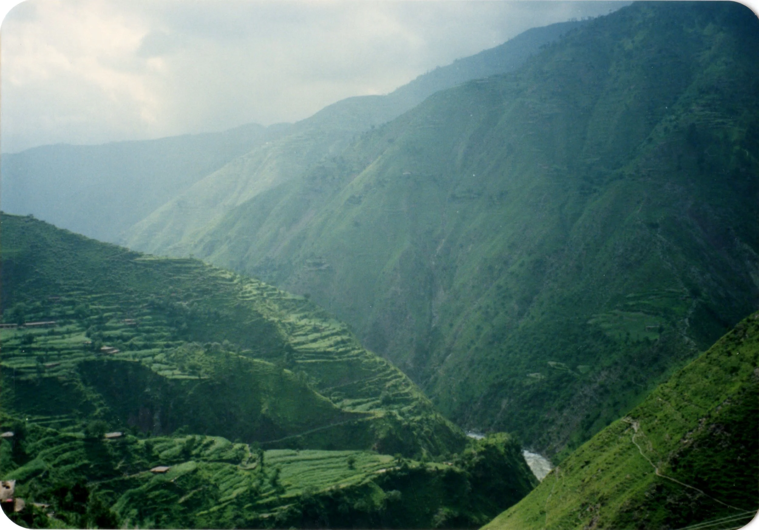 Lush green terraced fields on steep hillsides in a mountainous landscape with cloudy sky.
