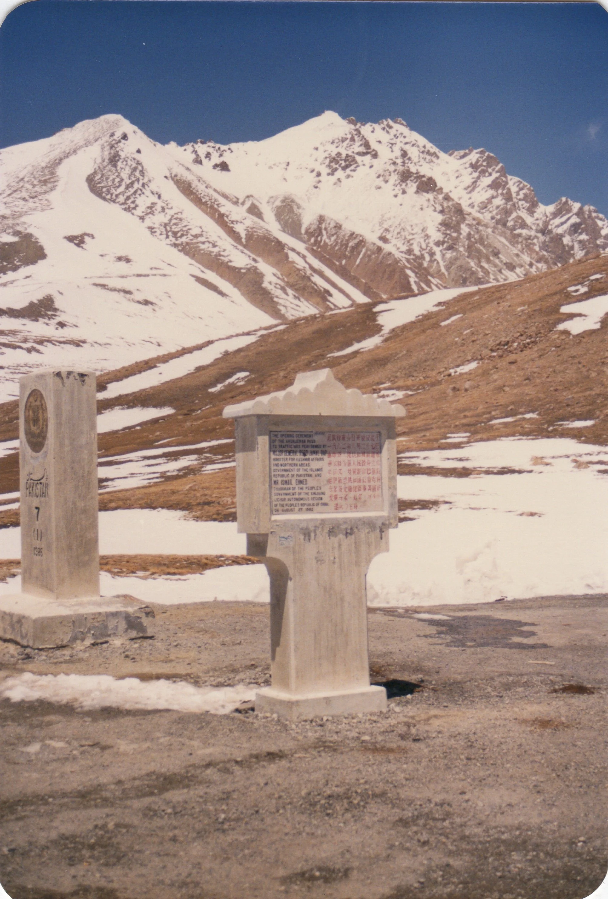 Snow-capped mountain range with a clear blue sky in the background. In the foreground, there are three concrete markers or monuments, one with Chinese writing and the others with text in English and numbers.