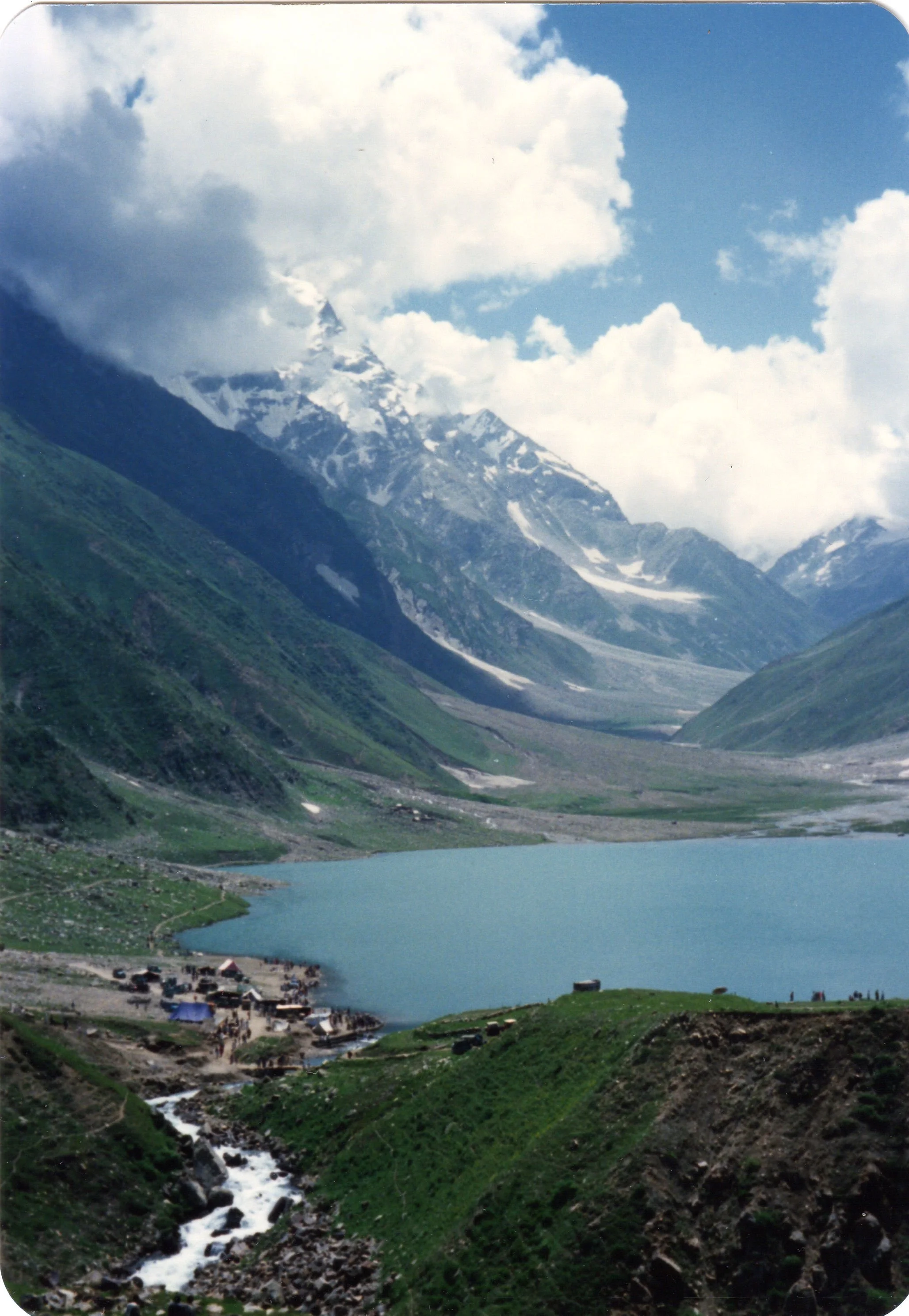 Mountain landscape with snow-capped peaks, a blue lake in the valley, and a small village near the water, green hills, and a river flowing down a rocky hillside.