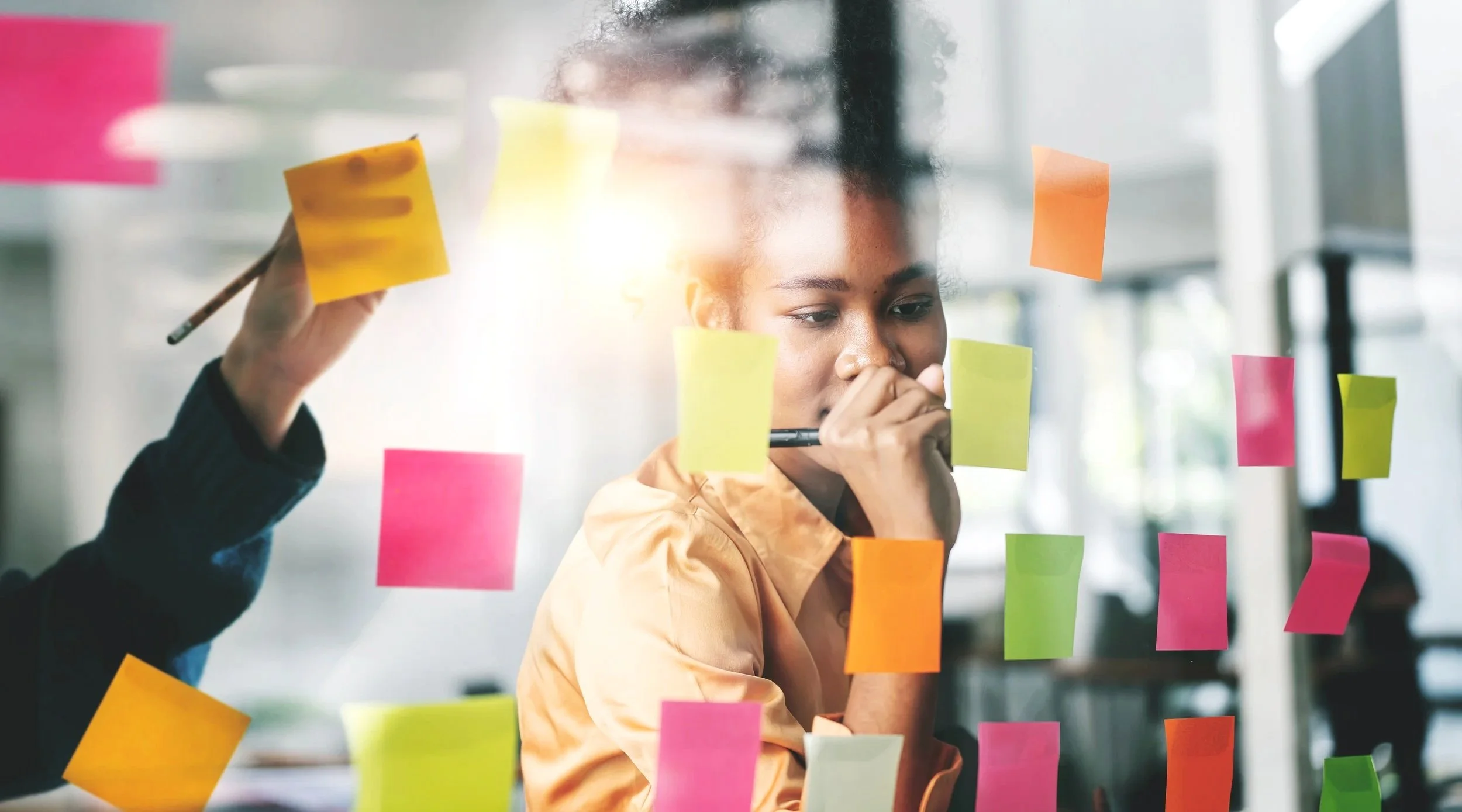 A woman with curly hair in a beige shirt is looking at colorful sticky notes on a glass wall or window while holding a pen to her face, in a bright office space.