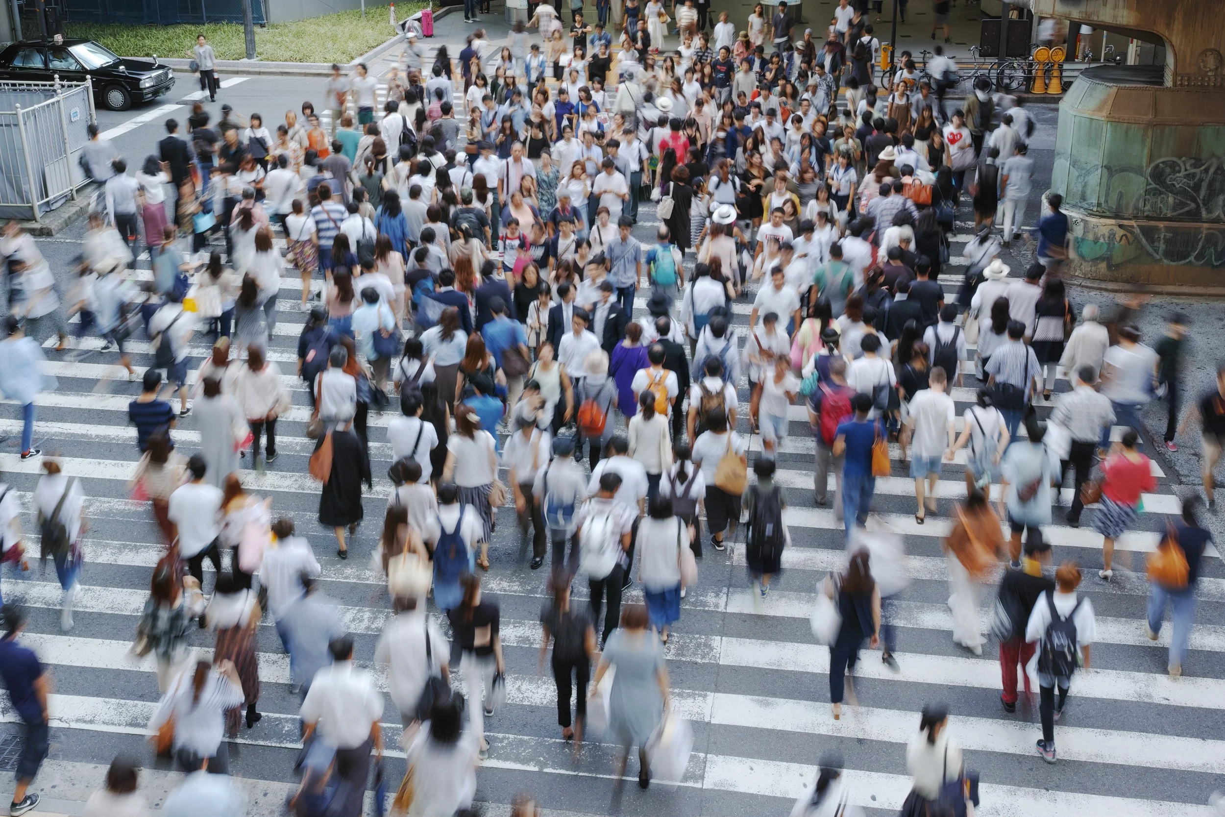 A crowded city street crosswalk with many pedestrians walking in various directions, some carrying bags and wearing hats. Vehicles are visible on the side of the street, and the background shows parts of buildings and graffiti-covered structures.