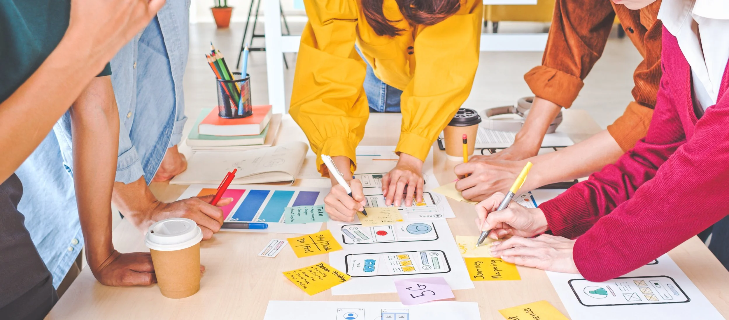 Group of people collaborating on a creative project at a table, with sketches, notes, and color swatches, in a bright, modern workspace.