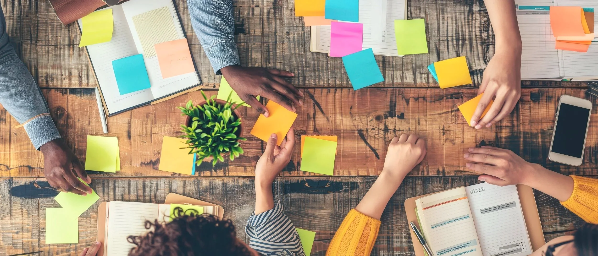 Top-down view of multiple hands working on a wooden table with notebooks, colorful sticky notes, a small potted plant, a smartphone, and pens, suggesting a collaborative work or brainstorming session.