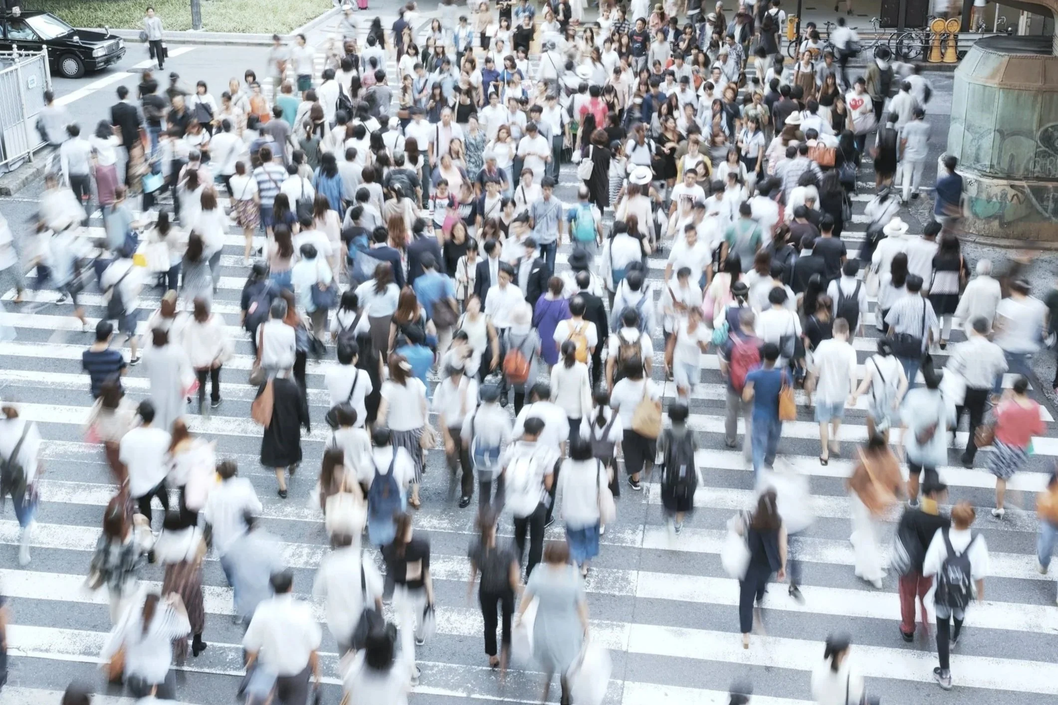 A crowded city crosswalk filled with pedestrians walking in various directions, with some blurred due to motion, near parked cars and urban buildings.