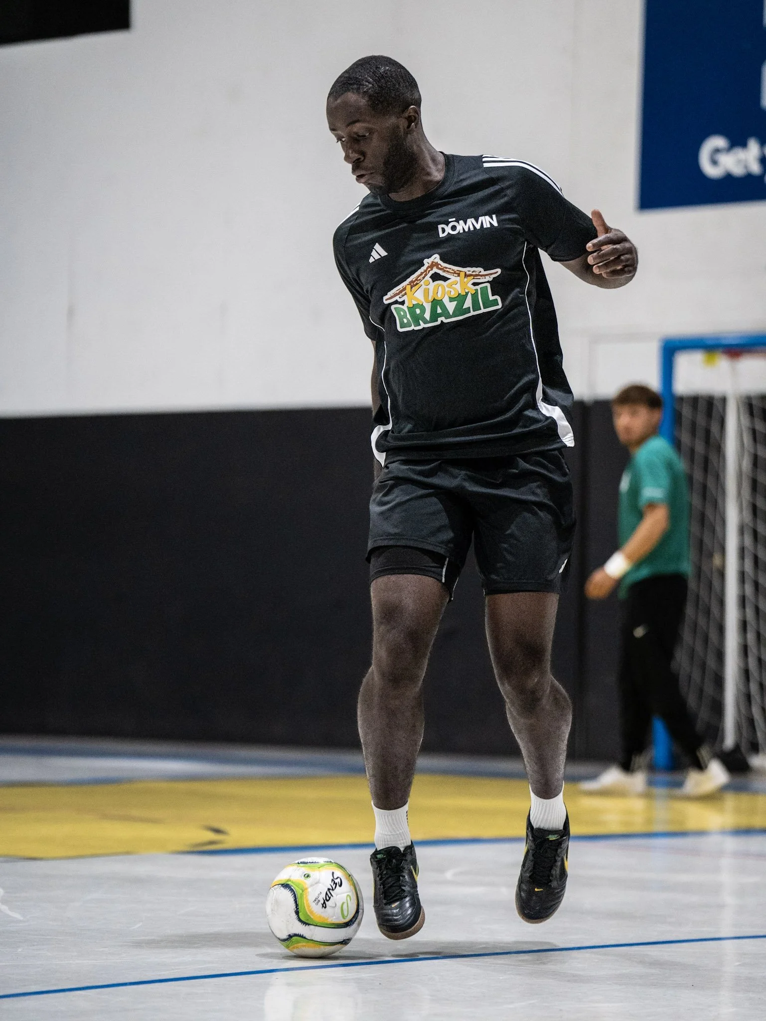 A male soccer player in black athletic clothing, with 'Kiosk Brazil' on his shirt, is dribbling a soccer ball on an indoor court, wearing black soccer shoes and white socks.
