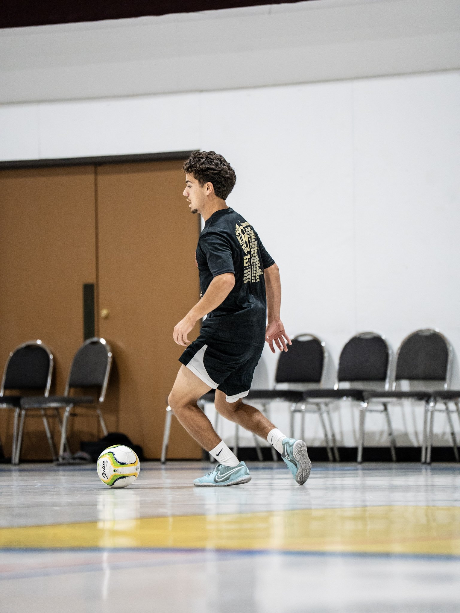 A young man in athletic clothing is playing indoor soccer, kicking a white and yellow soccer ball on a gym floor with chairs lined up in the background.