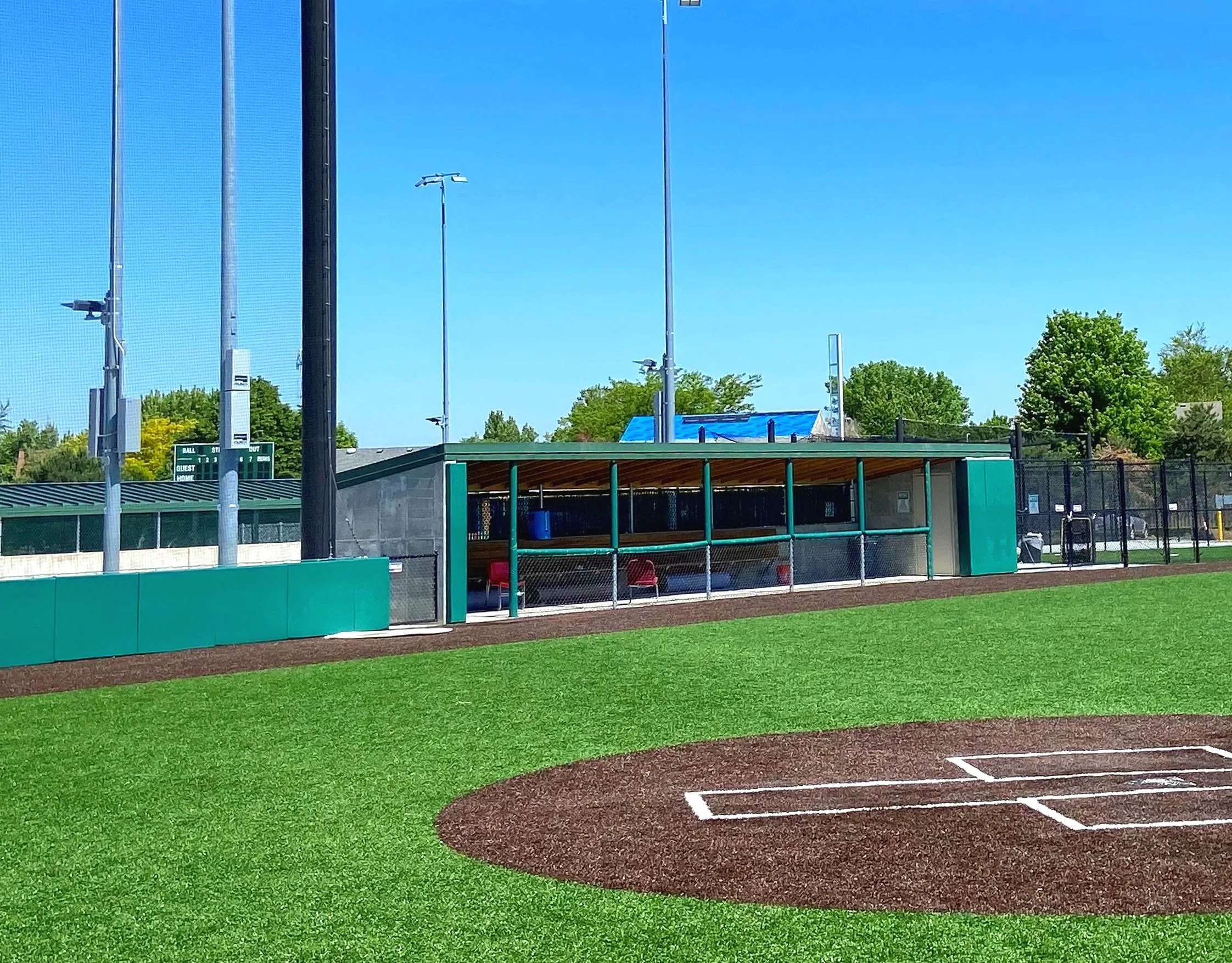 Teal green baseball dugout with dugout rail padding and integrated barrier netting system at professional synthetic turf baseball facility viewed from pitcher's mound — USS baseball dugout supplier and sports field padding supplier and sports field n