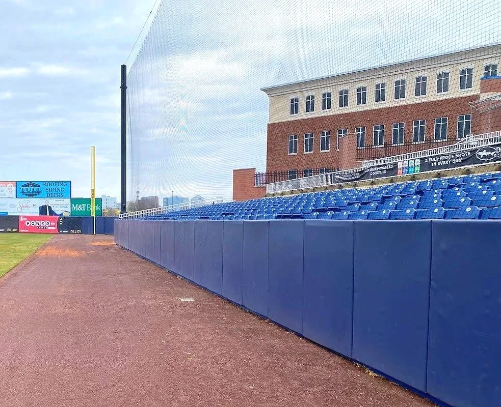 Navy blue exterior wall padding installed along full outfield fence line with overhead barrier netting system at professional level baseball facility with multi-level brick grandstand and press box — USS exterior wall padding supplier and sports fiel