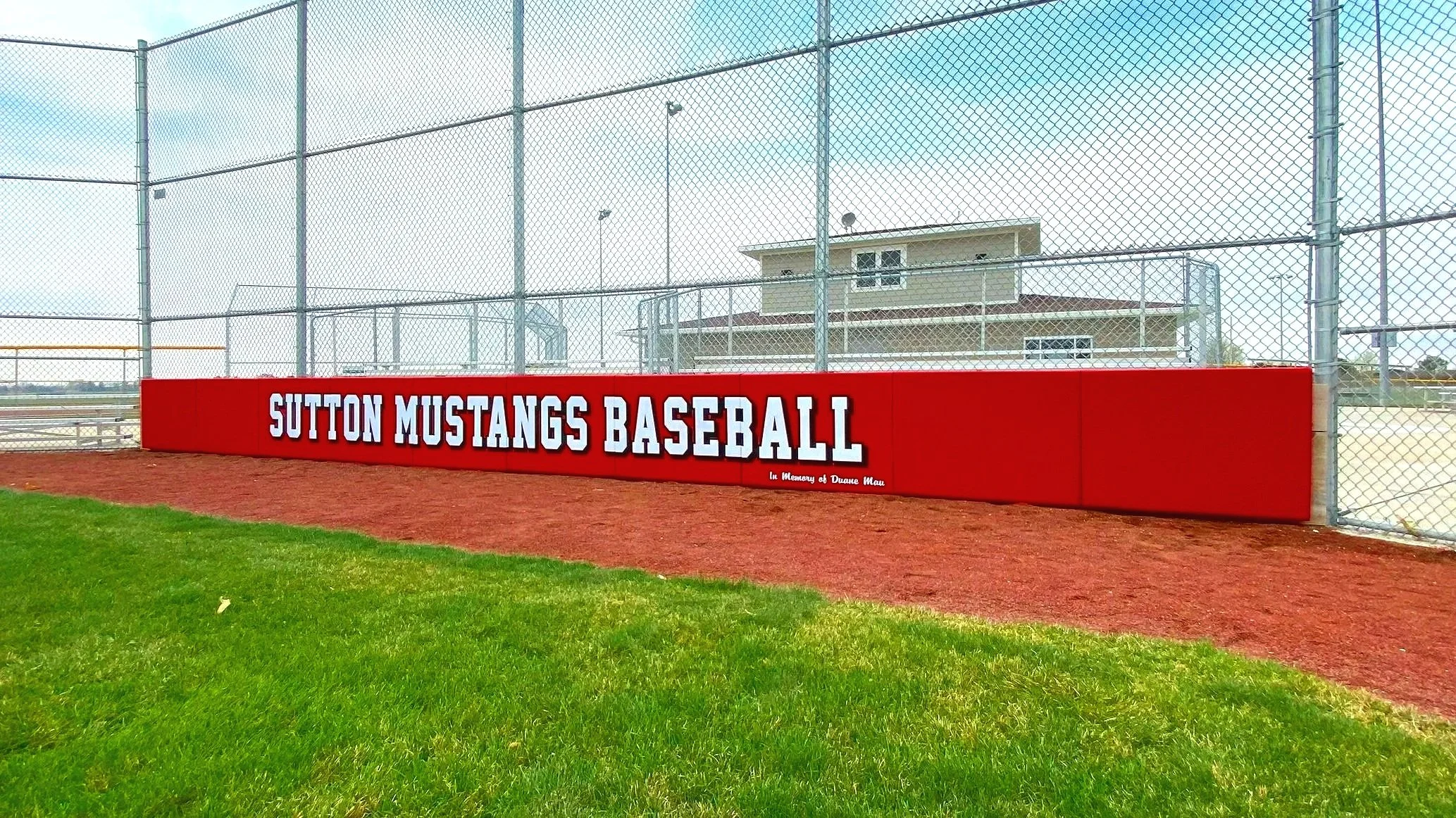 Custom branded red exterior wall padding displaying Sutton Mustangs Baseball team name along full outfield fence line at Sutton Nebraska athletic facility with press box — USS exterior wall padding supplier and athletic facility branding graphics sup