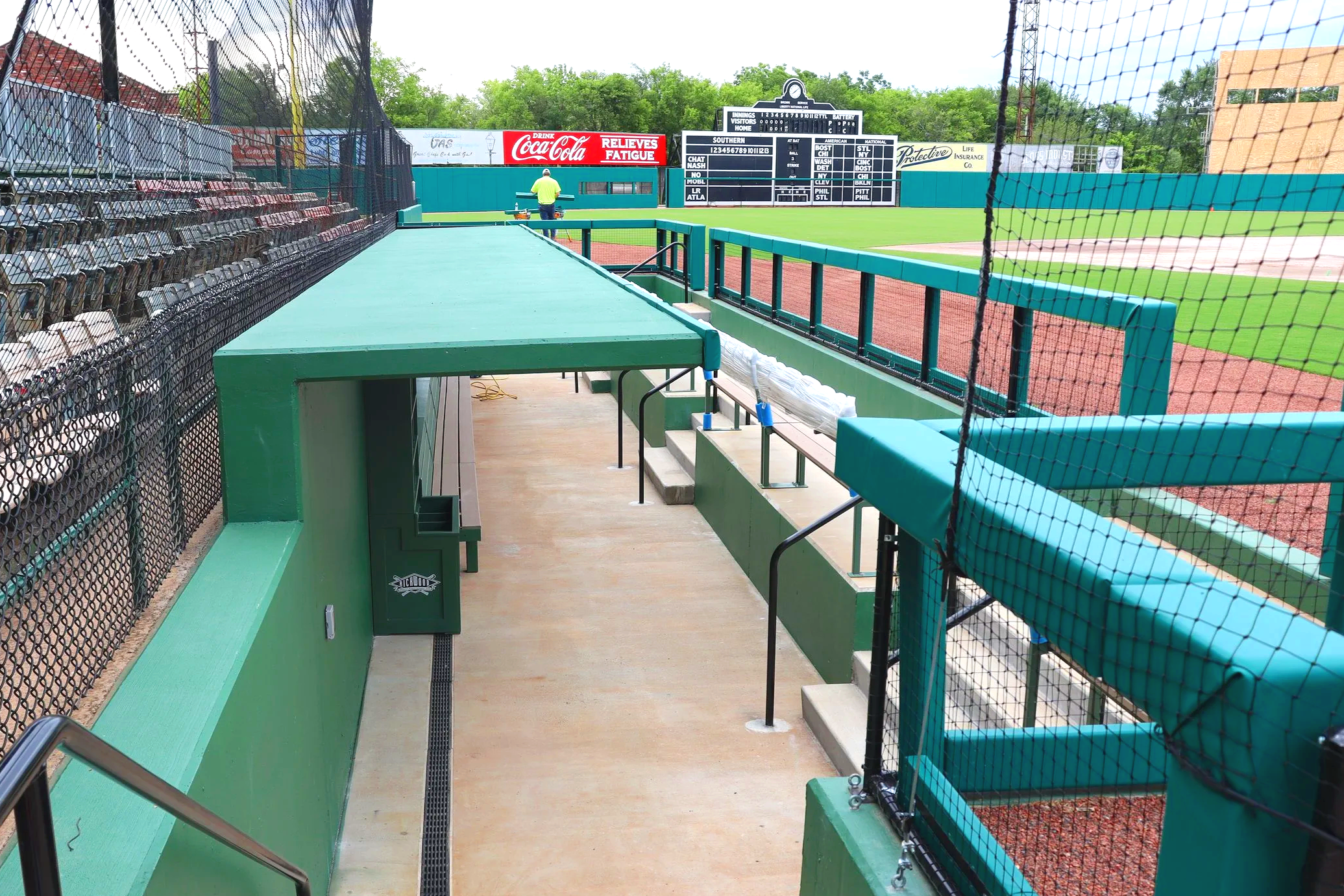 Teal green historic baseball dugout interior with dugout rail padding and barrier netting at Rickwood Field in Birmingham Alabama showing grandstand seating scoreboard and warning track — USS dugout rail padding supplier and sports field netting supp