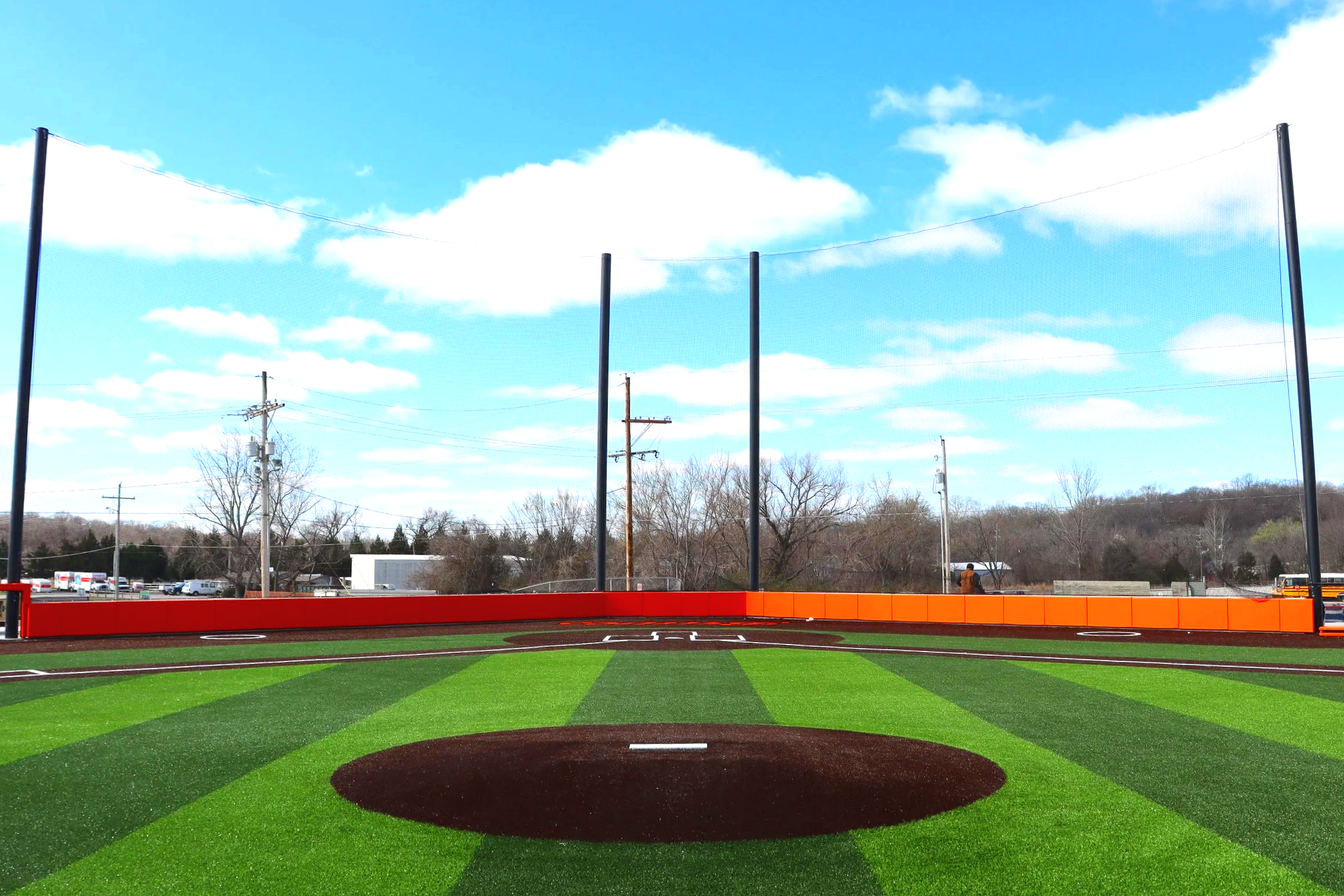 Orange and red custom branded exterior wall padding along full outfield fence line with barrier netting poles at Bonner Springs Kansas baseball facility viewed from pitcher's mound — USS exterior wall padding supplier and sports field padding supplie