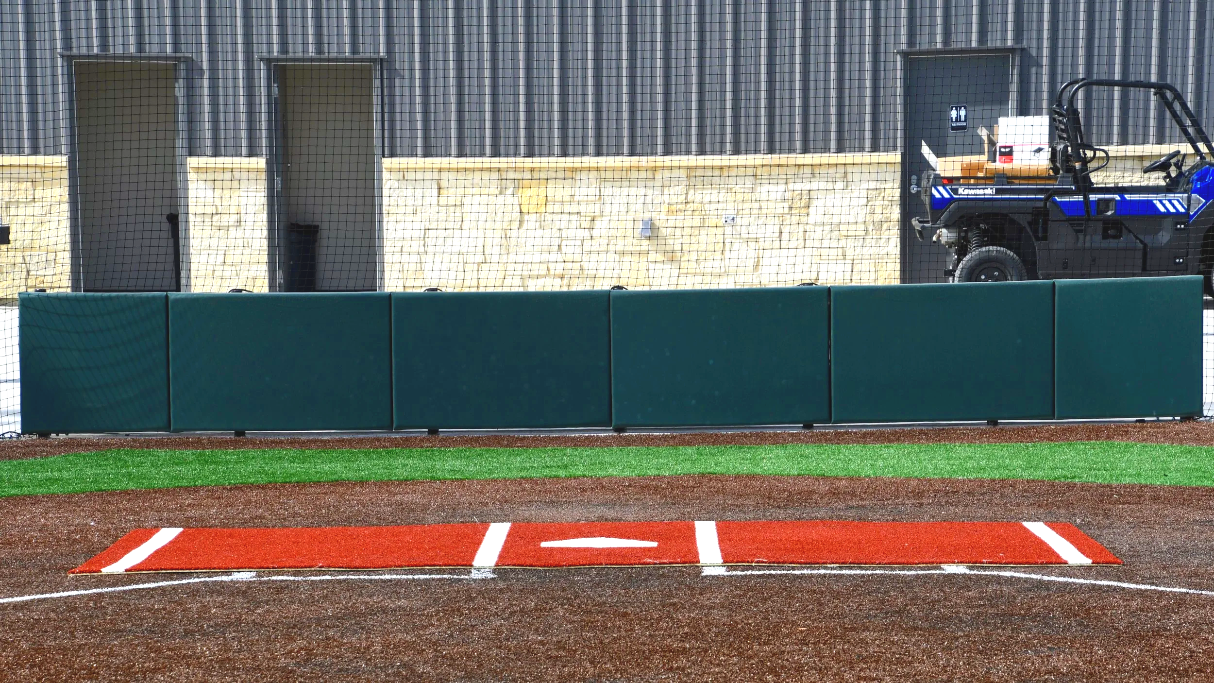 Dark green exterior wall padding installed along full facility building wall behind home plate area at professional baseball facility with red synthetic turf warning track — USS exterior wall padding supplier and sports field padding supplier providi