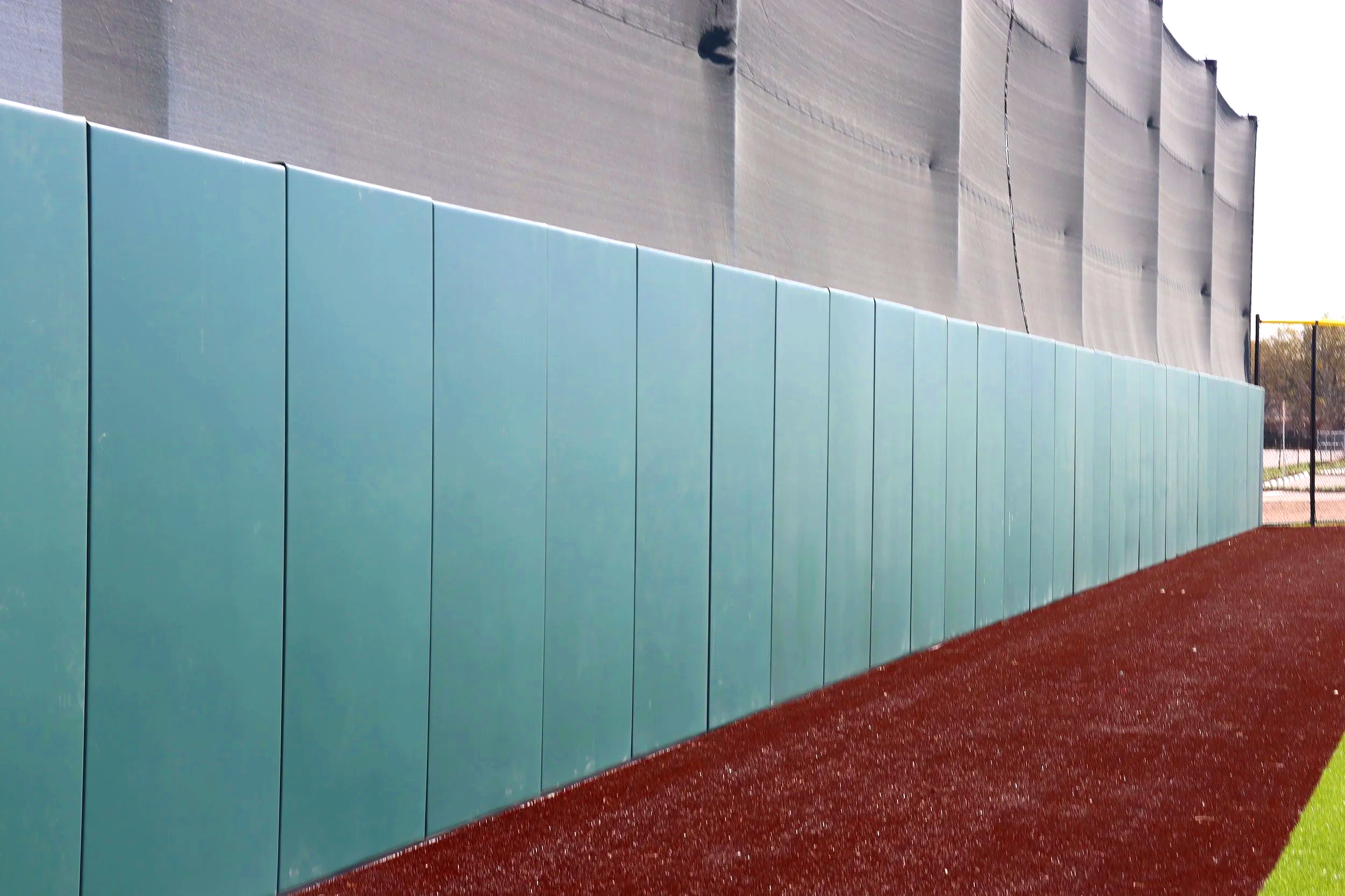 Teal exterior wall padding panels installed along full length of large scale athletic facility building exterior wall with red warning track — USS exterior wall padding supplier and sports field padding supplier providing complete wall to wall protec