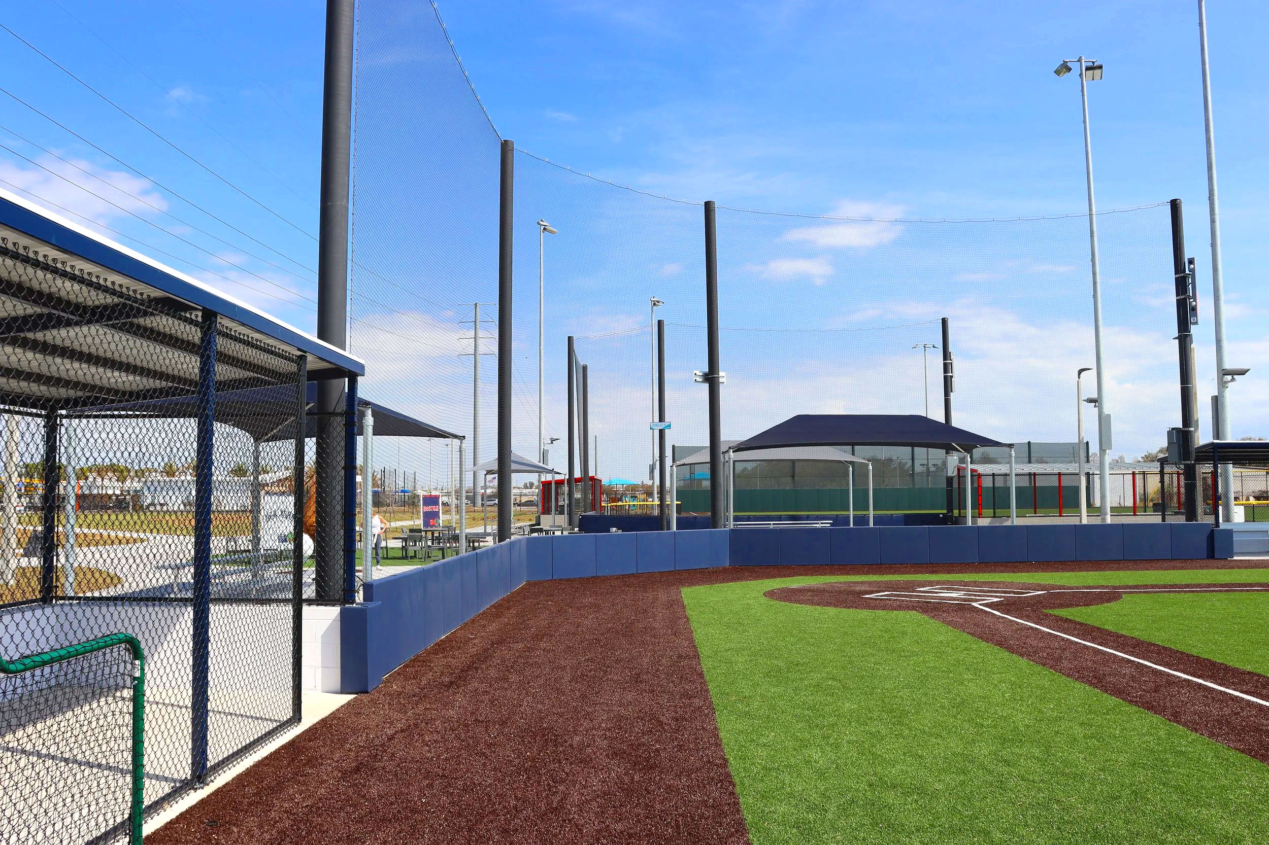 Blue exterior wall padding along full outfield and foul line fence with overhead barrier netting system and dugout barrier netting at Plummer Park baseball facility with synthetic turf and red warning track — USS exterior wall padding supplier and sp