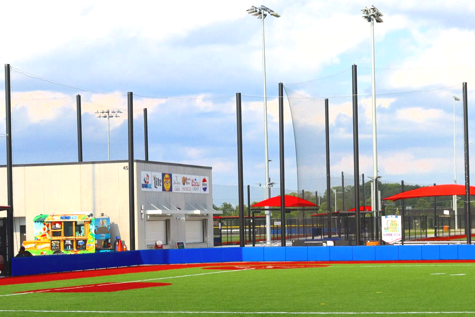 Blue exterior wall padding along full outfield fence line with red cantilever shade structures, foul poles, and barrier netting at professional baseball complex — USS sports field padding supplier