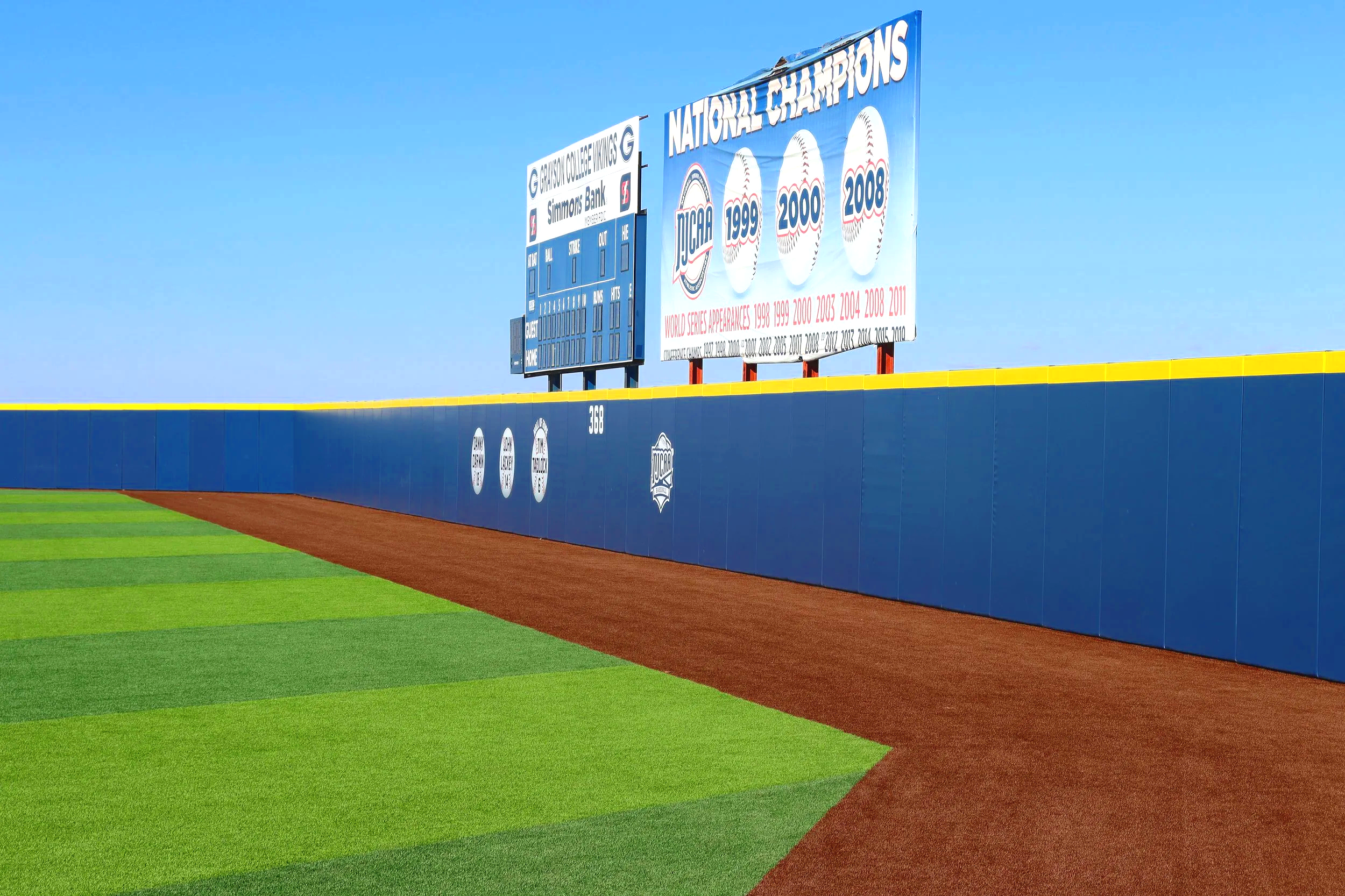 Blue exterior wall padding installed along the full outfield fence line at Grayson College baseball field — NCAA compliant sports field wall padding supplier USS