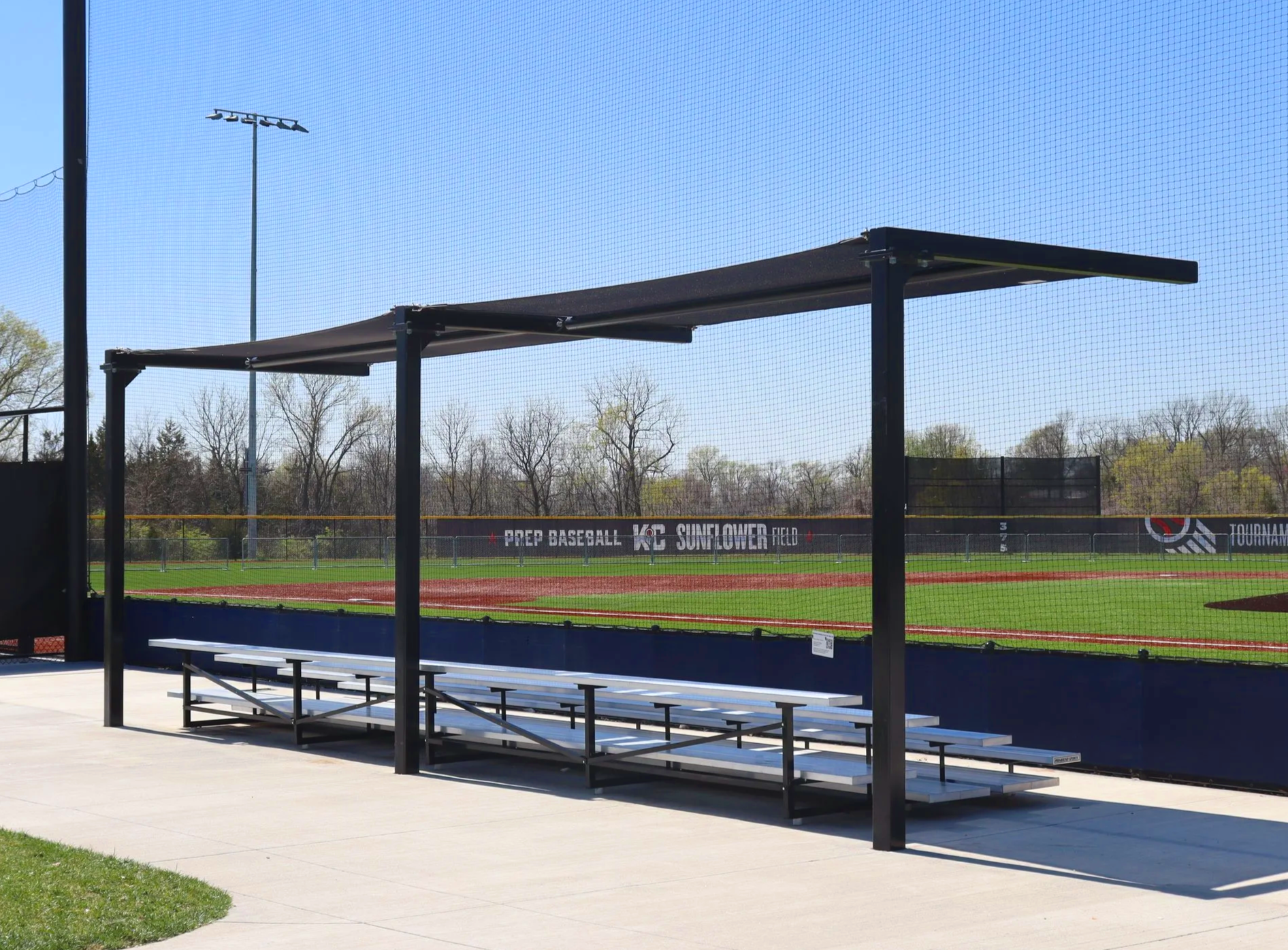 T-Post shade structure over aluminum bleacher seating at Homefield Kansas City baseball facility with branded windscreen — USS shade structures for athletic fields supplier