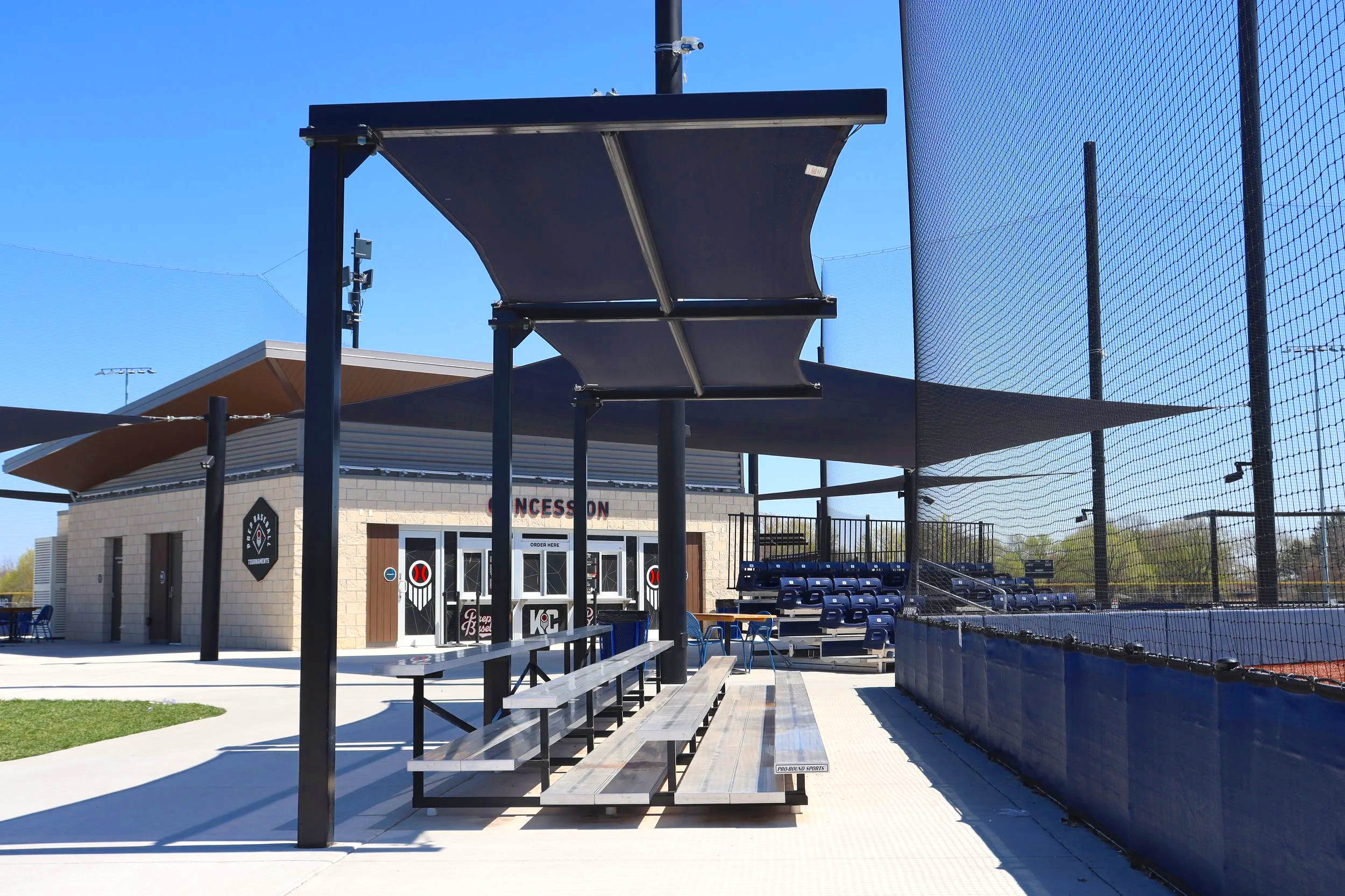 Hip shade structure over spectator benches and concession area at Homefield Kansas City athletic complex with barrier netting and bleacher seating — USS shade structures for athletic fields supplier