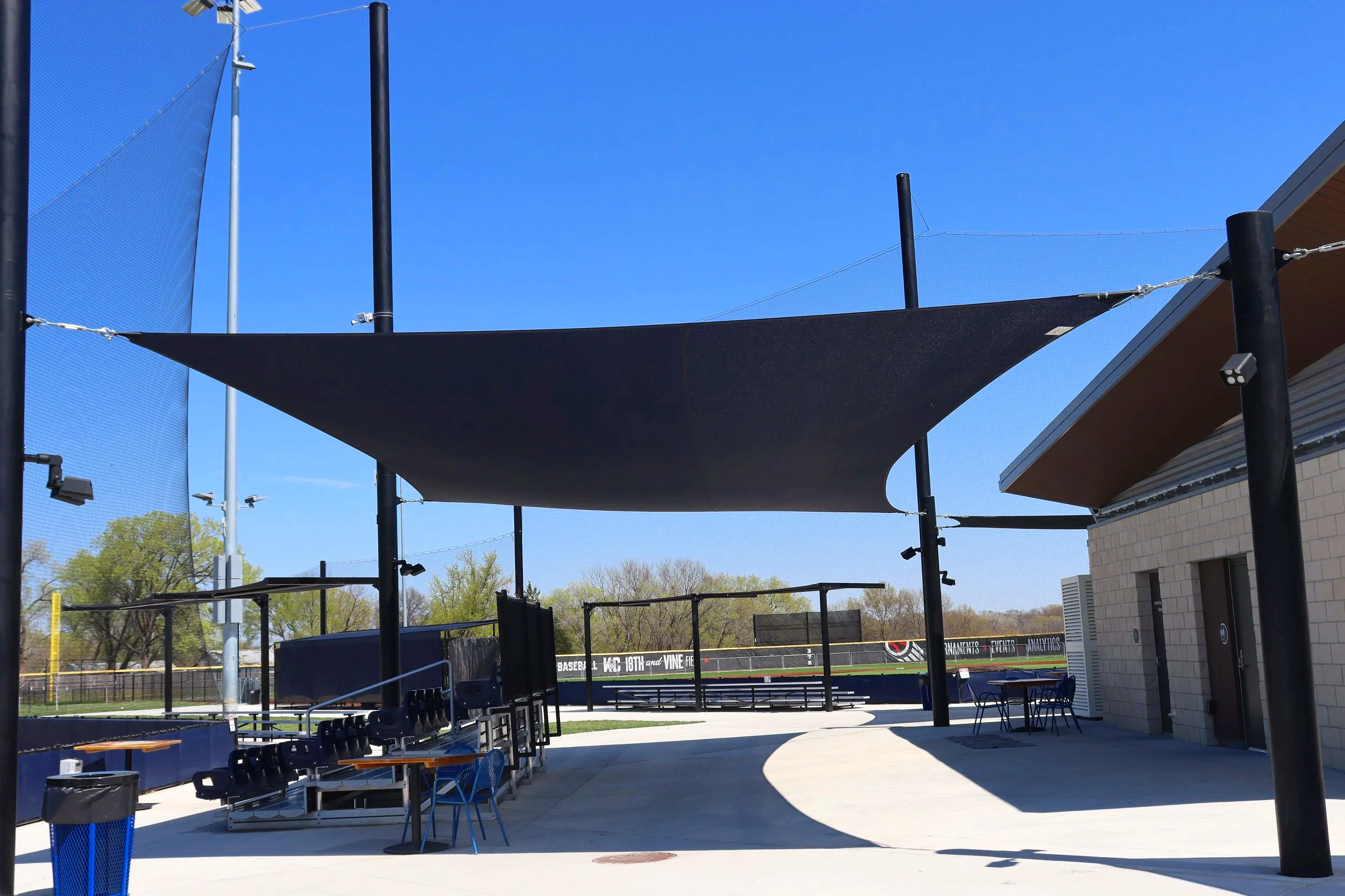 Sail shade structure over athletic complex common area and spectator gathering space at Homefield Kansas City with barrier netting and bleacher seating — USS shade structures for athletic fields supplier