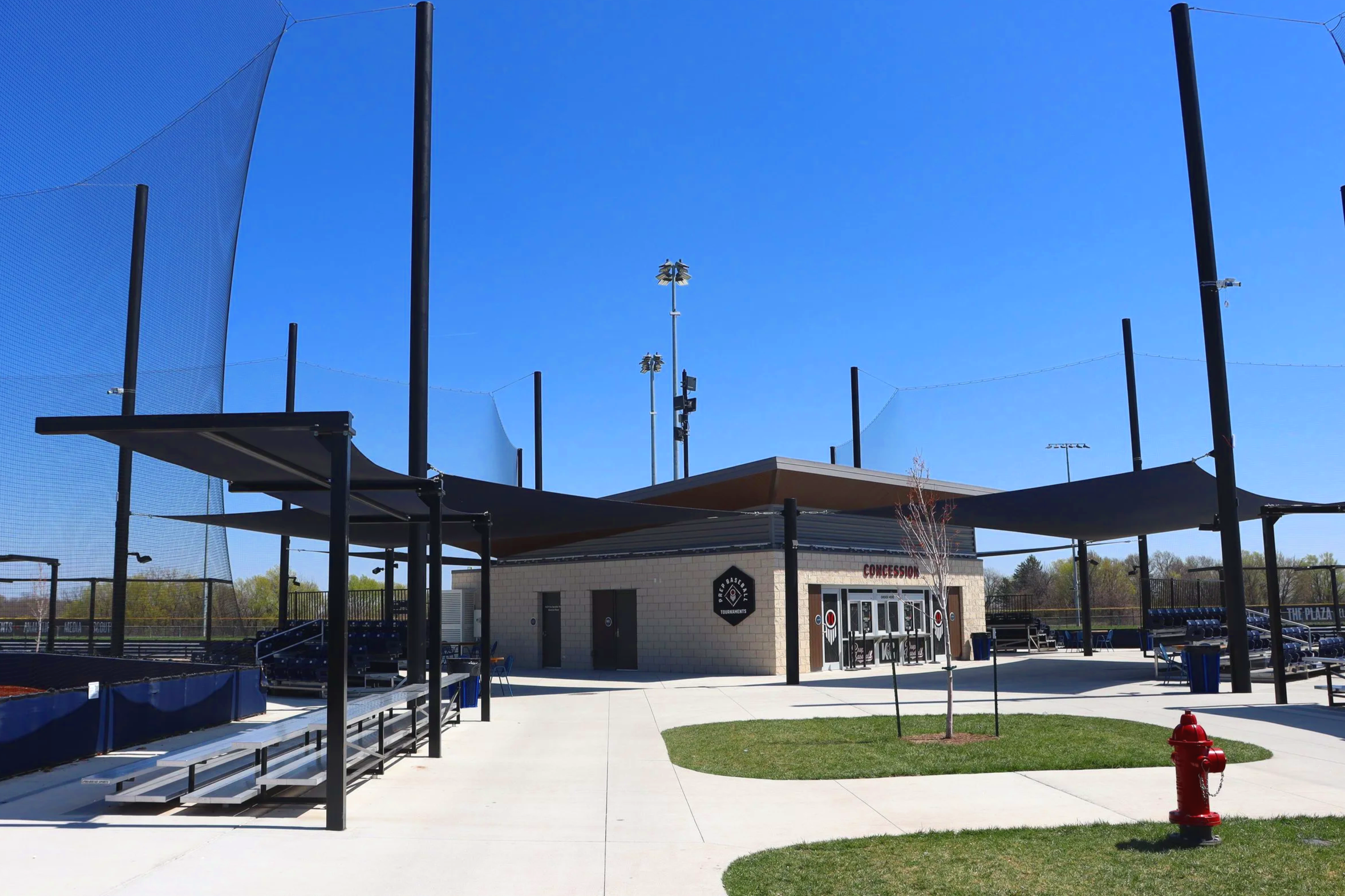 Cantilever shade structures over athletic complex common area and bleacher seating at Homefield in Kansas City with barrier netting and concession building — USS shade structures for athletic fields supplier