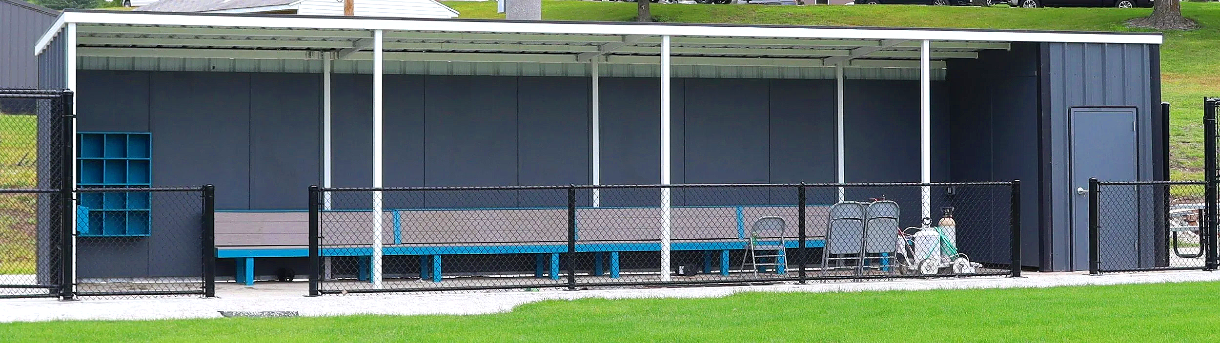 Fielder's Choice PRO Plus baseball dugout by USS in Corning, Iowa, with white steel roof frame, gray panel walls, teal-accented player benches, built-in helmet cubbies, and attached storage room.