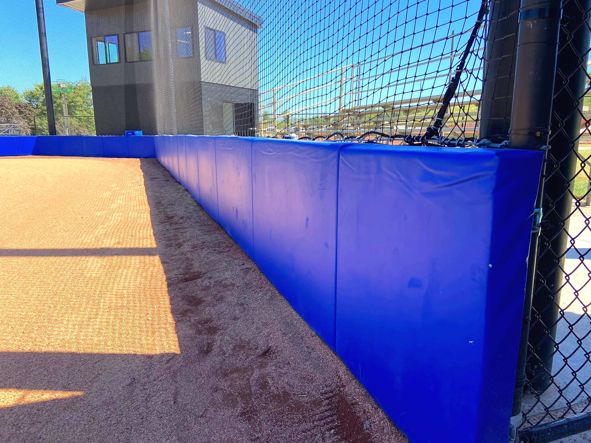 Blue exterior wall padding installed along chain link fence line with warning track and barrier netting overhead — USS sports field padding supplier