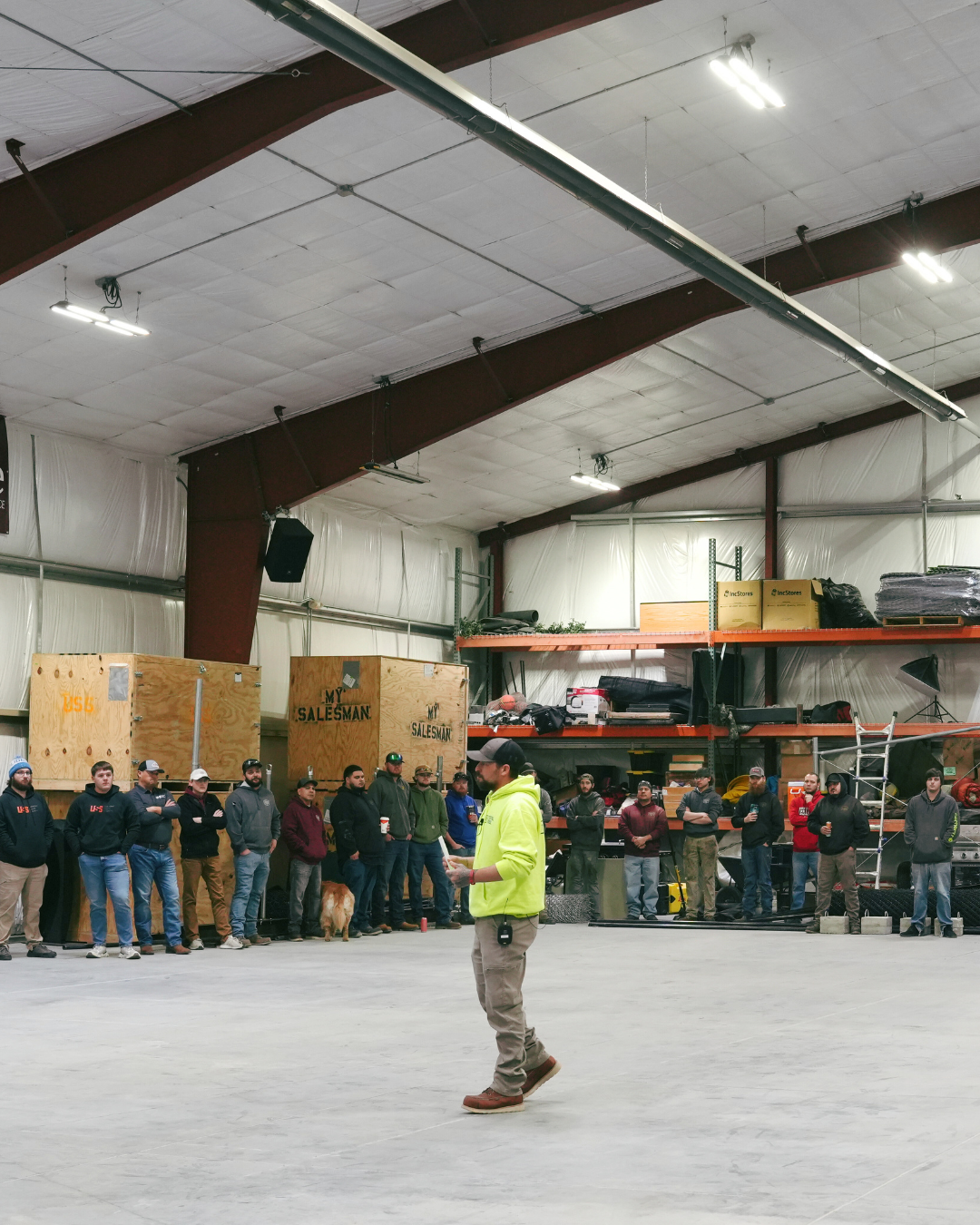 Crown Co. team members gathered in warehouse facility for company meeting led by team leader in yellow jacket