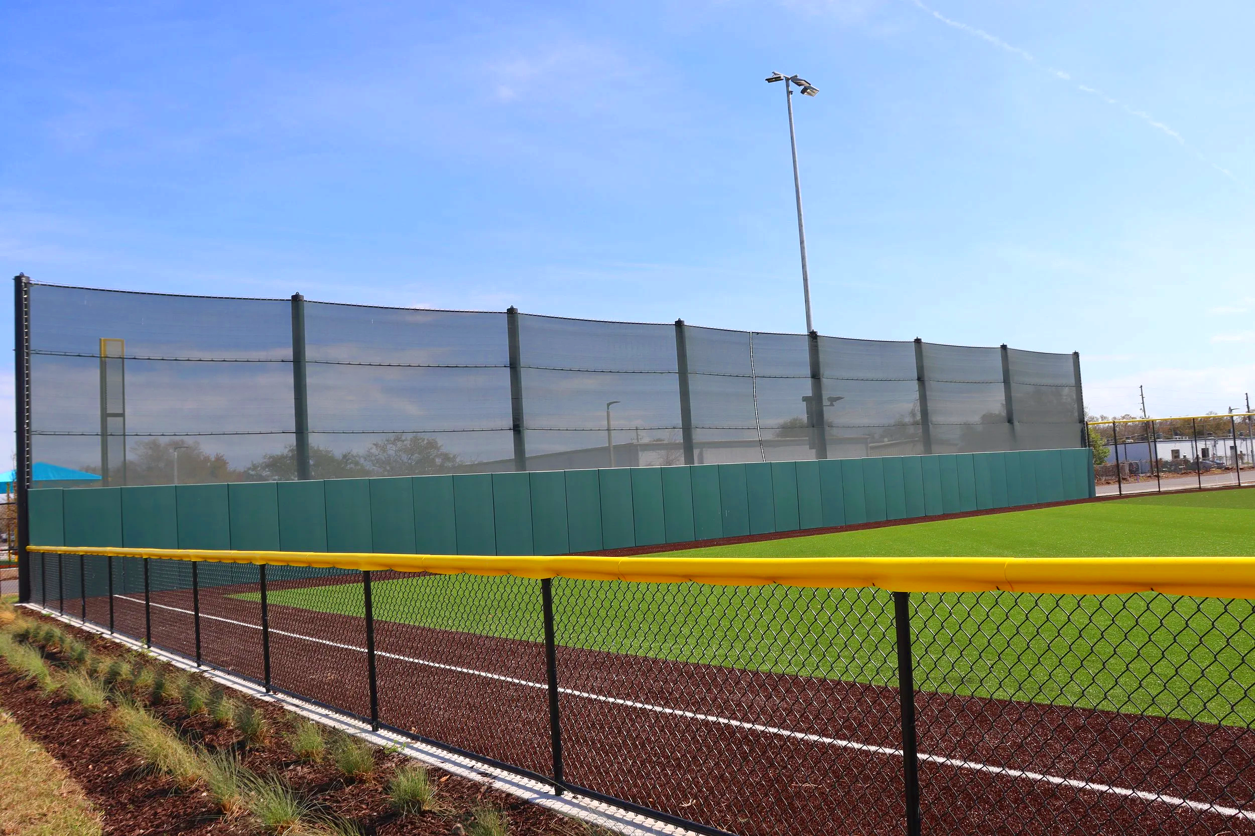 Yellow fence cap on chain link outfield fence at professional baseball complex with green wall padding and windscreen