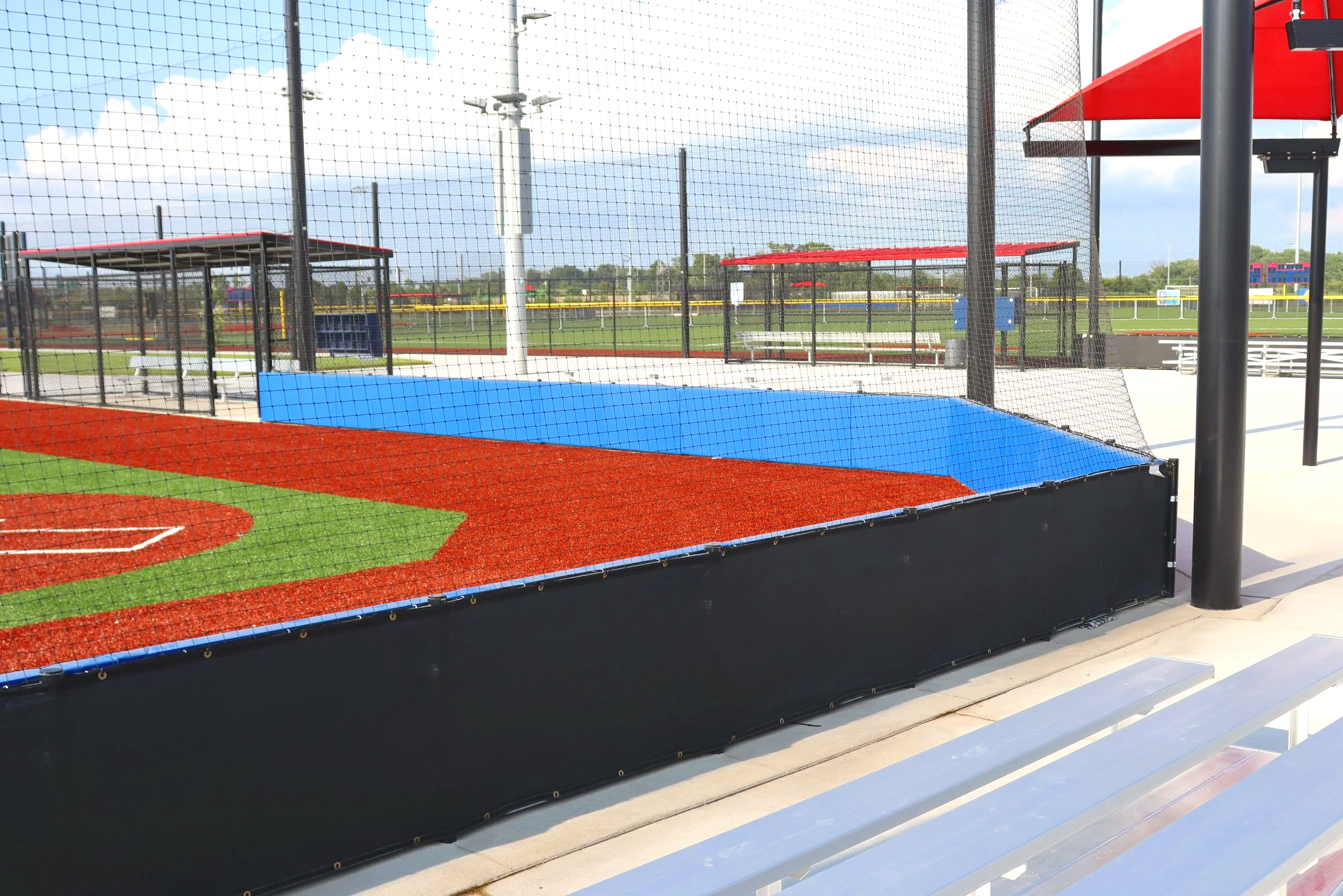 Black exterior wall padding installed along synthetic turf baseball field infield wall with aluminum bleachers and barrier netting overhead — USS sports field padding supplier