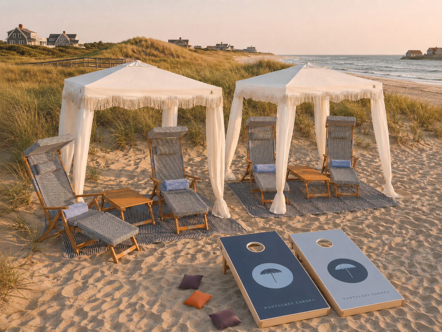 Beach scene with two cabanas, lounge chairs, and cornhole boards near the shoreline with houses on dunes in the background.