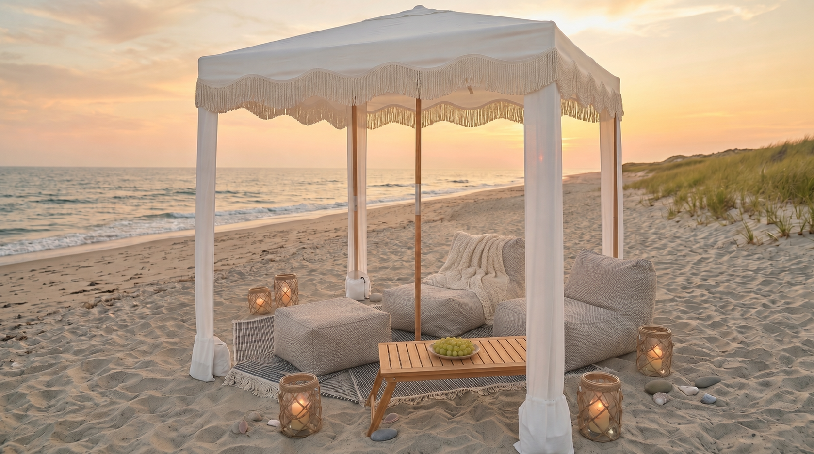 A cozy outdoor beach cabana with a canopy, pillows, and a small table with grapes, surrounded by lanterns with candles, at sunset on the beach.