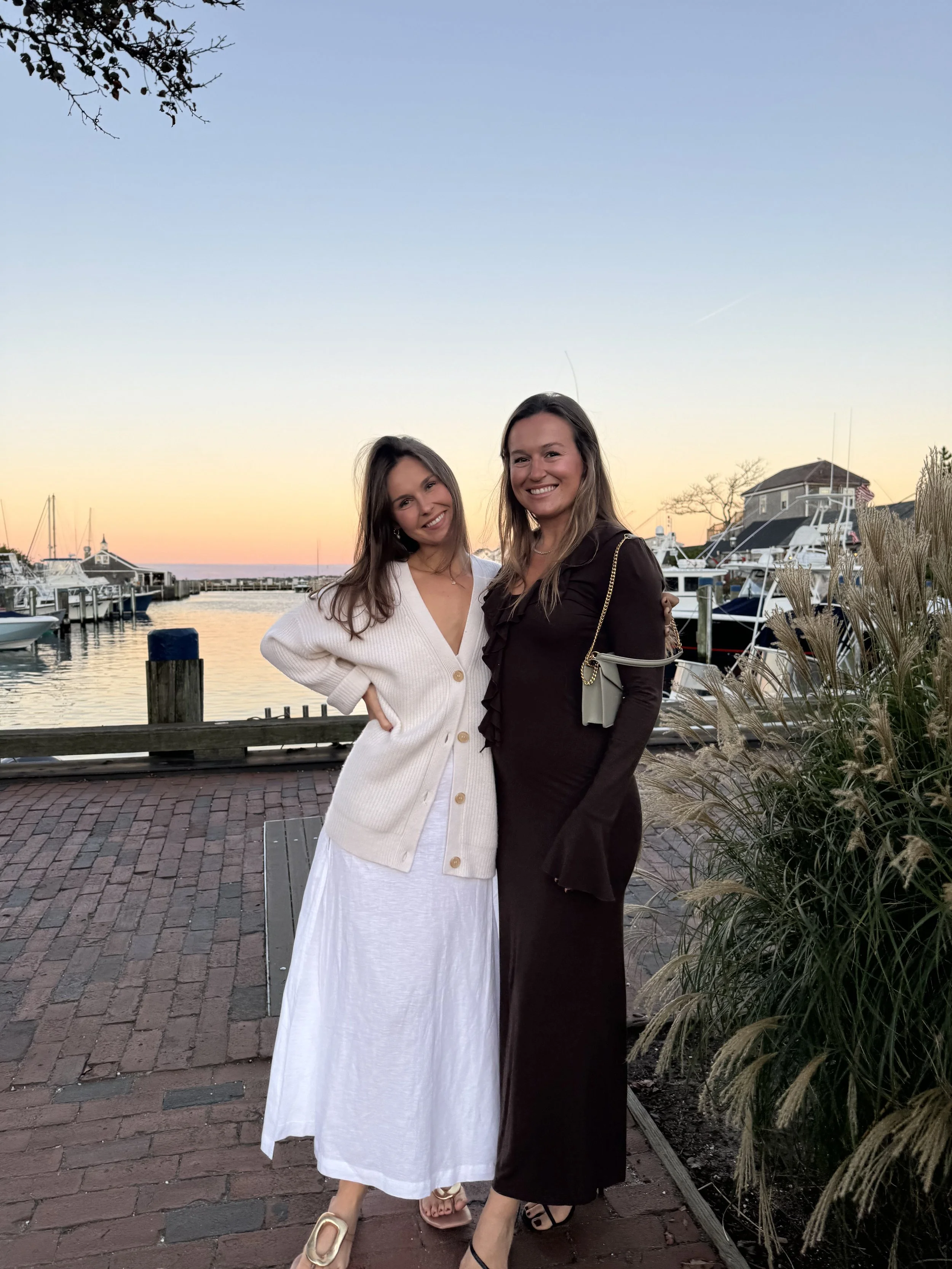 Two women standing together by a Nantucket marina at sunset, smiling for the camera. One is wearing a white dress with a button-up cardigan and sandals, the other is wearing a dark dress with black strappy heels and carrying a beige purse.