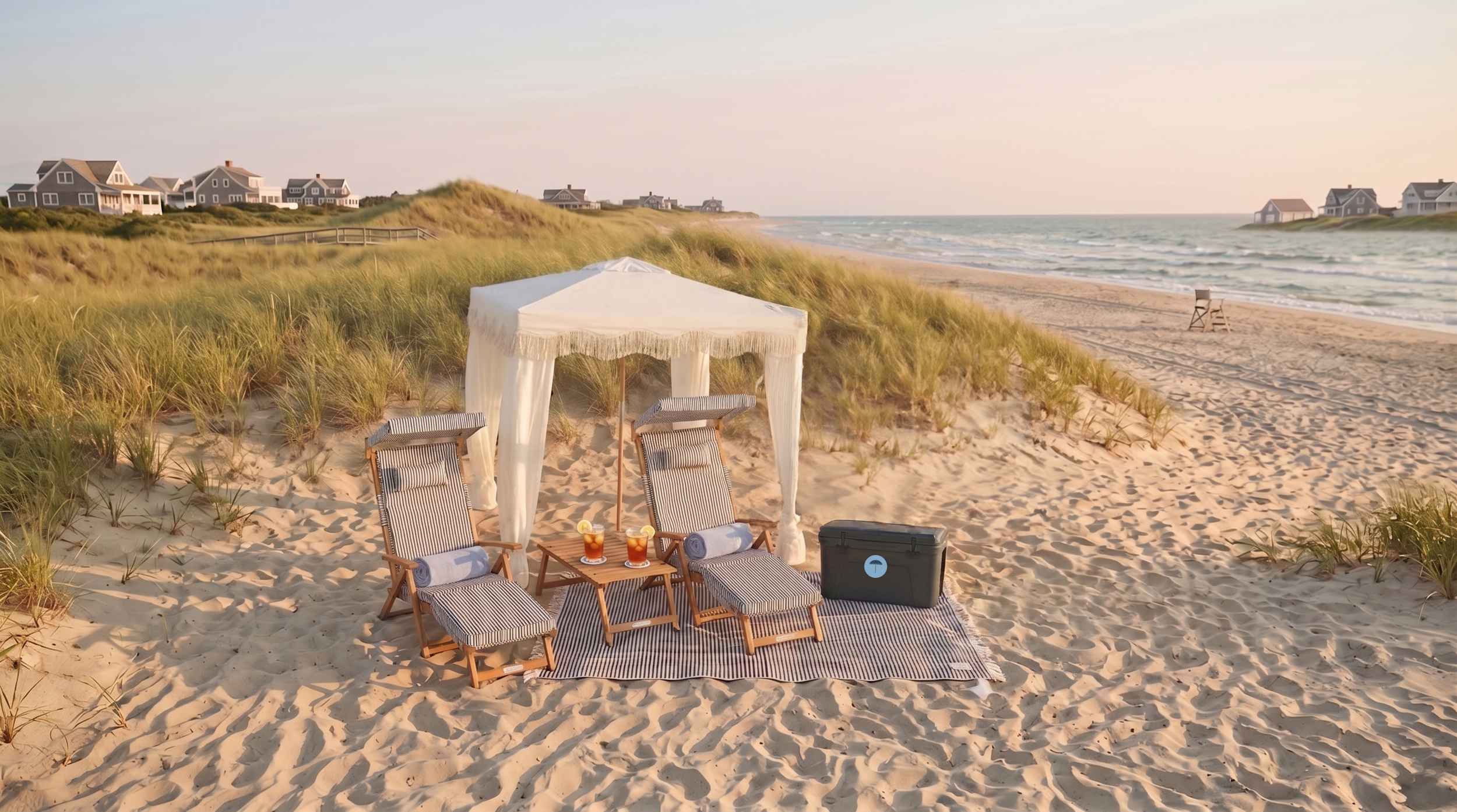 Beach scene with sand, grassy dunes, a white cabana with two striped lounge chairs, a small table with two drinks, and houses in the background near the shoreline of a Nantucket beach.