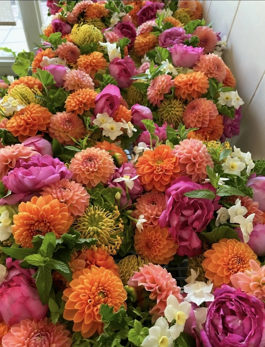 Colorful bouquet of various flowers including orange dahlias, pink peonies, white primroses, and yellow spider mums arranged on a surface near a window.