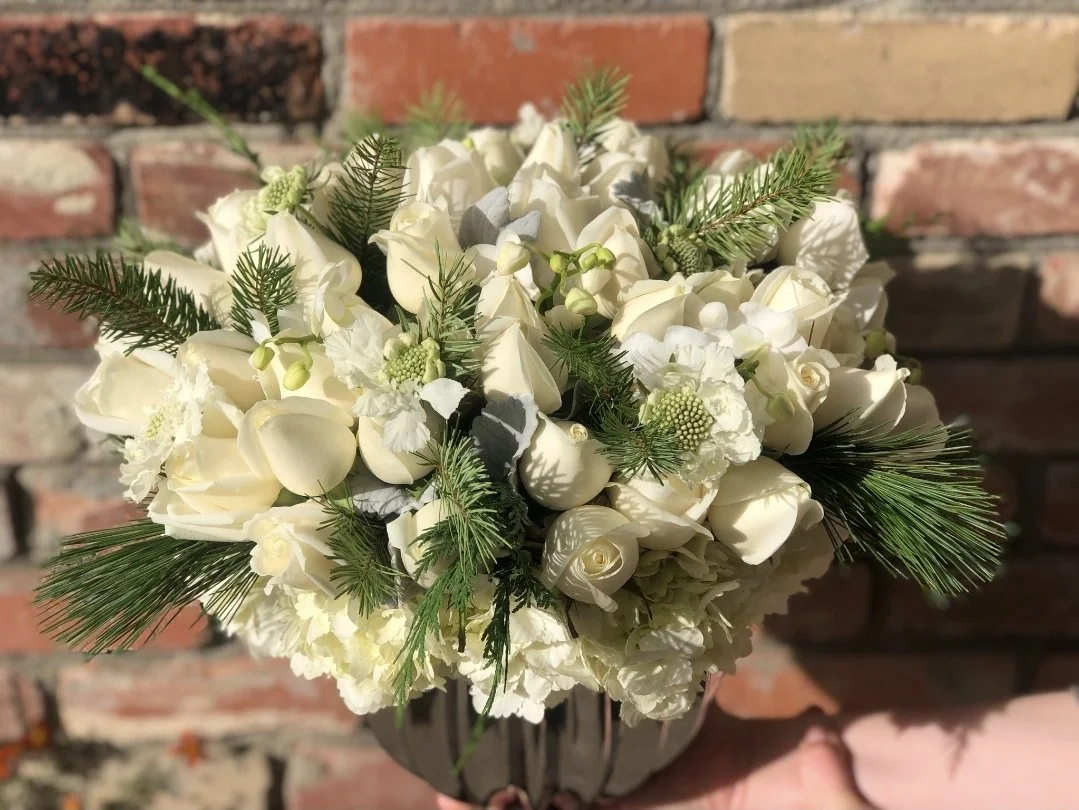 A bouquet of white roses, hydrangeas, and other flowers with greenery, held against a brick wall background.