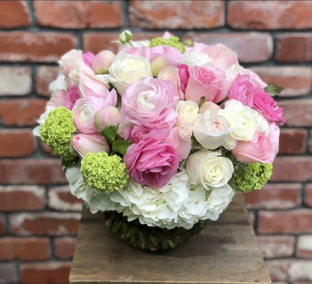 A round bouquet of pink, white, and green flowers, including roses, hydrangeas, and ranunculus, placed on a wooden surface with a brick wall in the background.