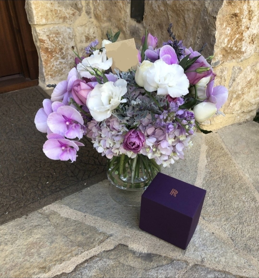 A bouquet of pink, white, and purple flowers in a glass vase, placed on a stone surface outside, next to a purple box with gold initials.