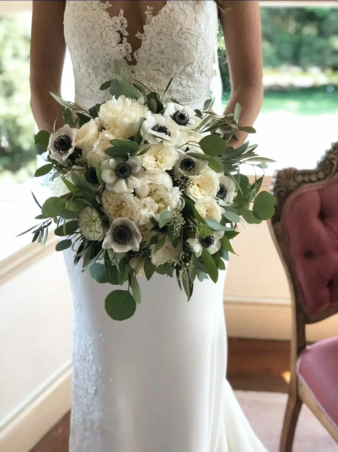 Bride holding a bouquet of white roses, anemones, and greenery, standing in a room with vintage pink chairs and a view of greenery outside.