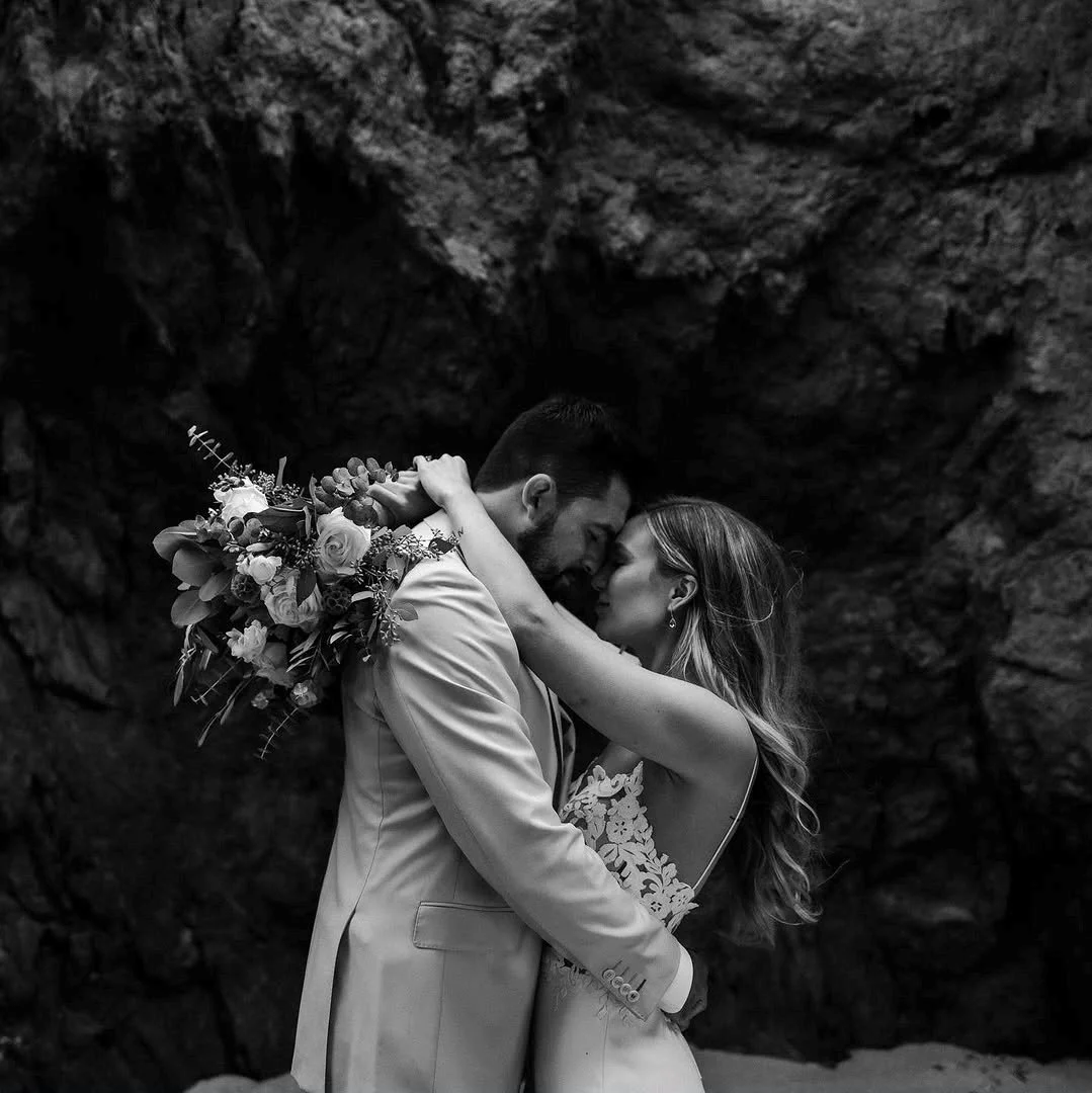 A black and white photo of a bride and groom embracing, with the groom holding a bouquet of flowers. They are touching foreheads and appear to be in a rocky outdoor setting.
