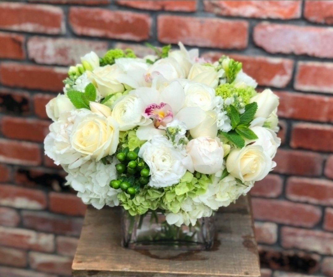 A bouquet of white roses, orchids, and green hydrangeas in a glass vase on a wooden surface, with a brick wall background.