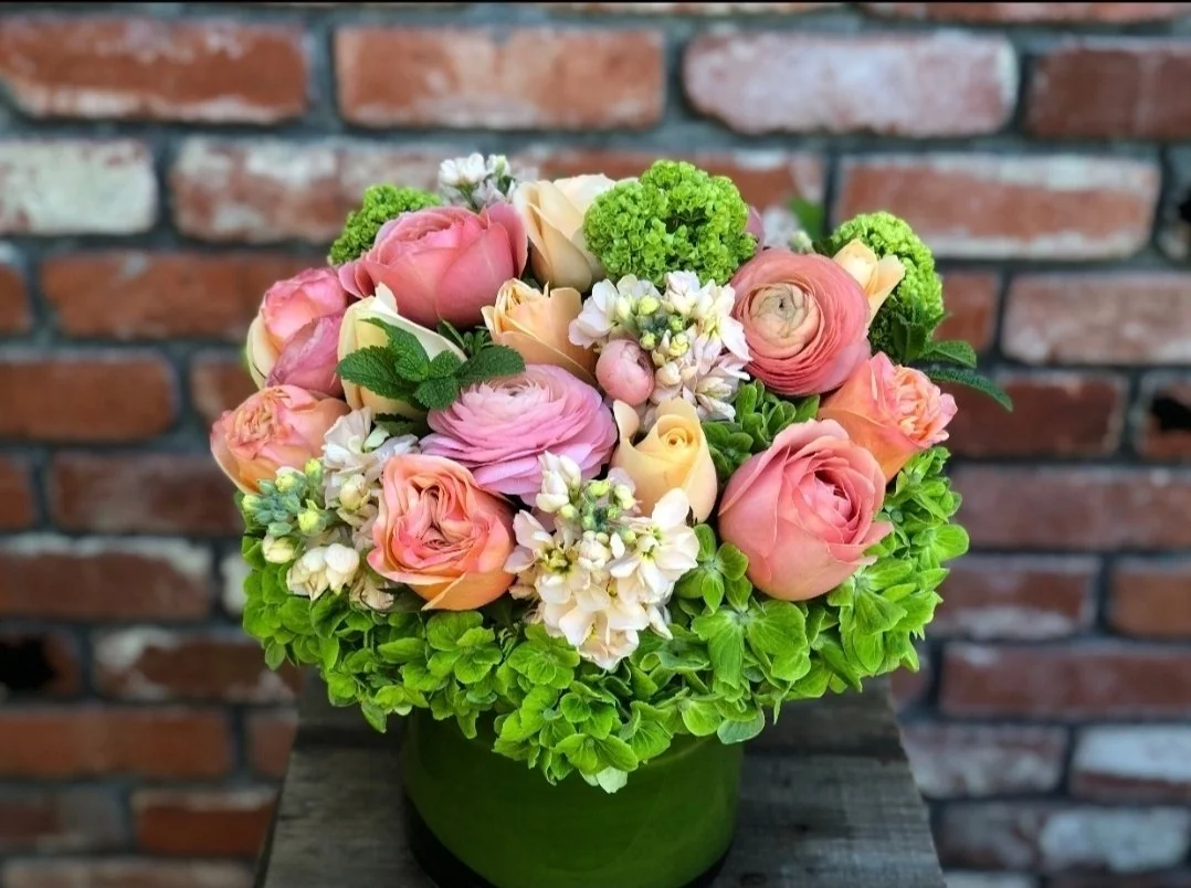 Pink and cream roses, ranunculus, small white flowers, green hydrangeas, and mint leaves arranged in a green vase, with a brick wall background.
